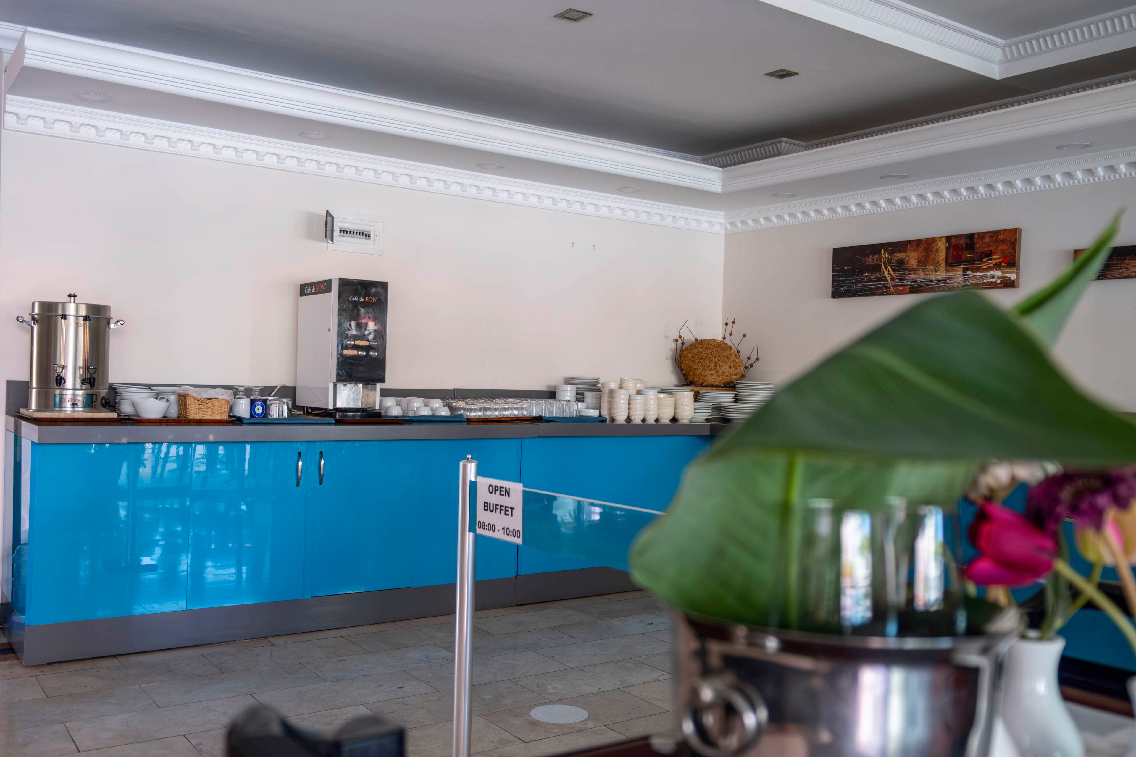 A hotel breakfast buffet counter features a bright blue cabinet with various dishes and cups neatly arranged. A large metal coffee urn and a beverage dispenser stand on the countertop, ready for guests. In the foreground, a metal ice bucket with green leaves and flowers adds a decorative touch. A small sign on a post reads "OPEN BUFFET" with additional text below, indicating service hours.