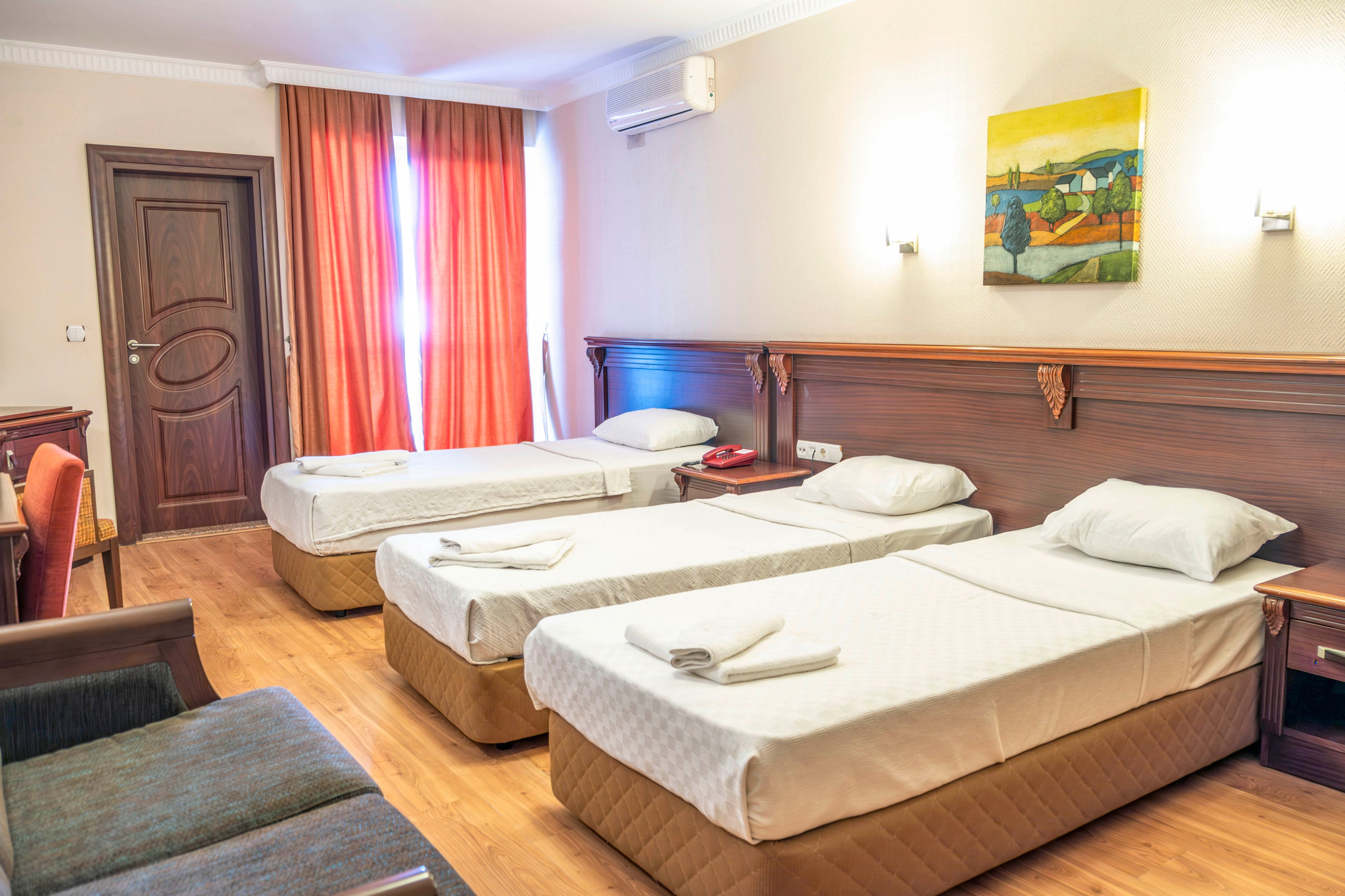A tidy hotel room features three single beds neatly made with white linens and folded towels. Warm wooden flooring and a wooden headboard run along the wall, complemented by a small desk and chair. Sunlight filters through red-orange curtains covering a large window, creating a cozy atmosphere. A colorful abstract painting and soft wall lighting add decorative touches to the simple, comfortable space.