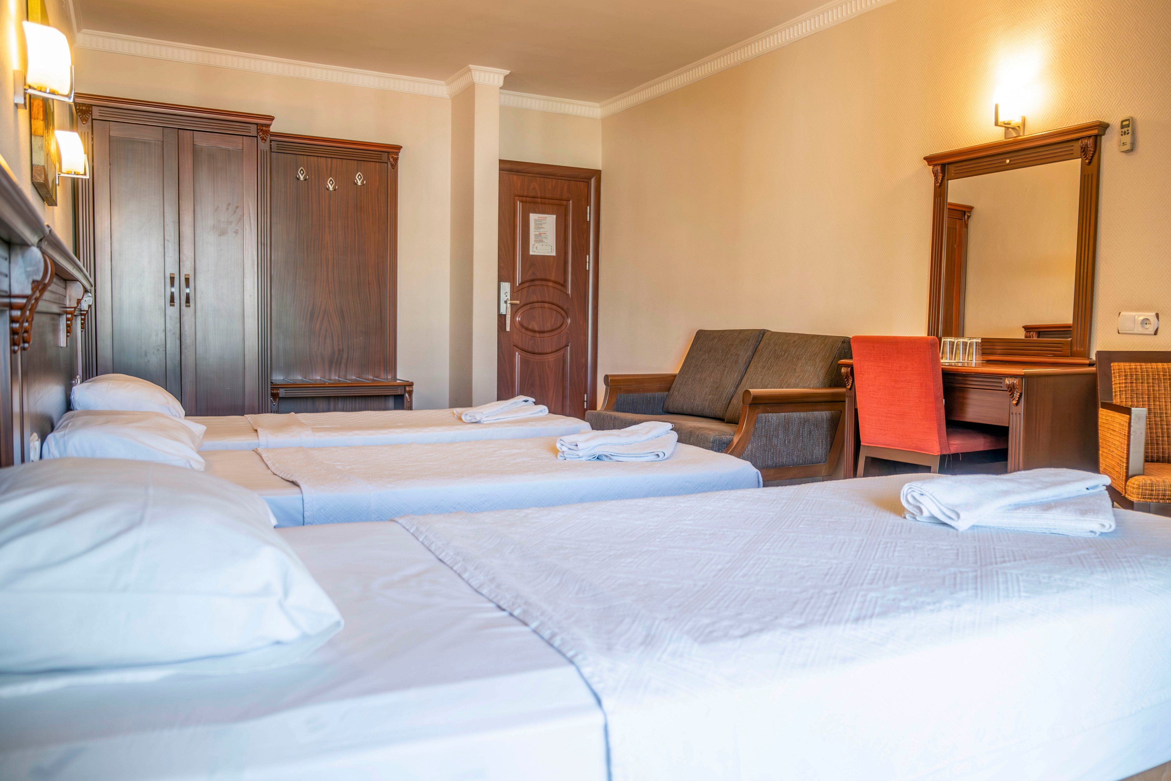 A tidy hotel room features three neatly made single beds with white linens in the foreground. The space includes wooden furniture such as a wardrobe, door, and a dressing table with a mirror. A small seating area with a sofa and an orange chair adds comfort to the room. Warm wall lighting and neutral colors create a cozy, inviting atmosphere suitable for guests.