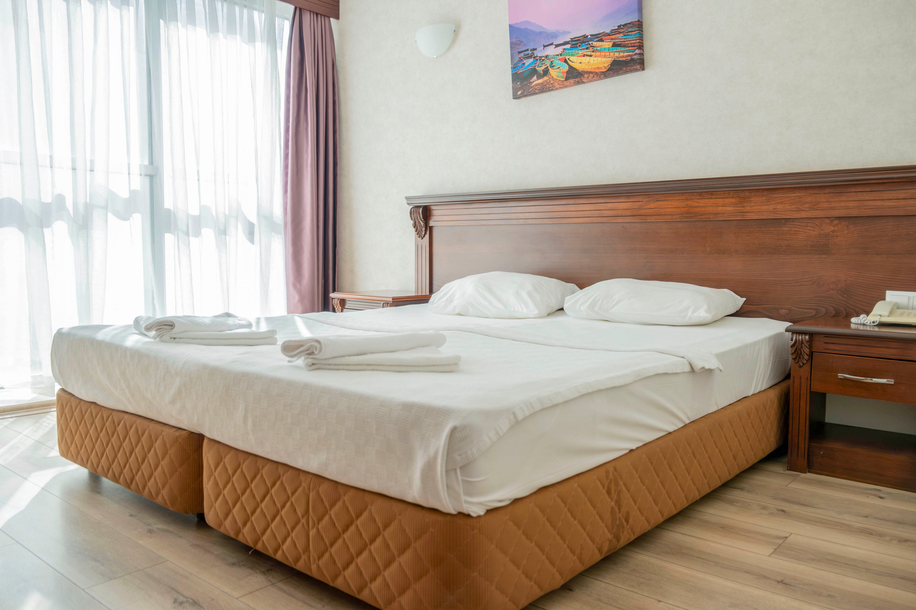 A neatly made double bed with white linens and two pillows stands in a bright hotel room. Sunlight filters through large sheer curtains beside the bed, creating a soft, inviting atmosphere. A wooden headboard, bedside table, and wall-mounted artwork add warmth and character to the space. The overall scene suggests a clean, comfortable place for rest or vacation accommodation.