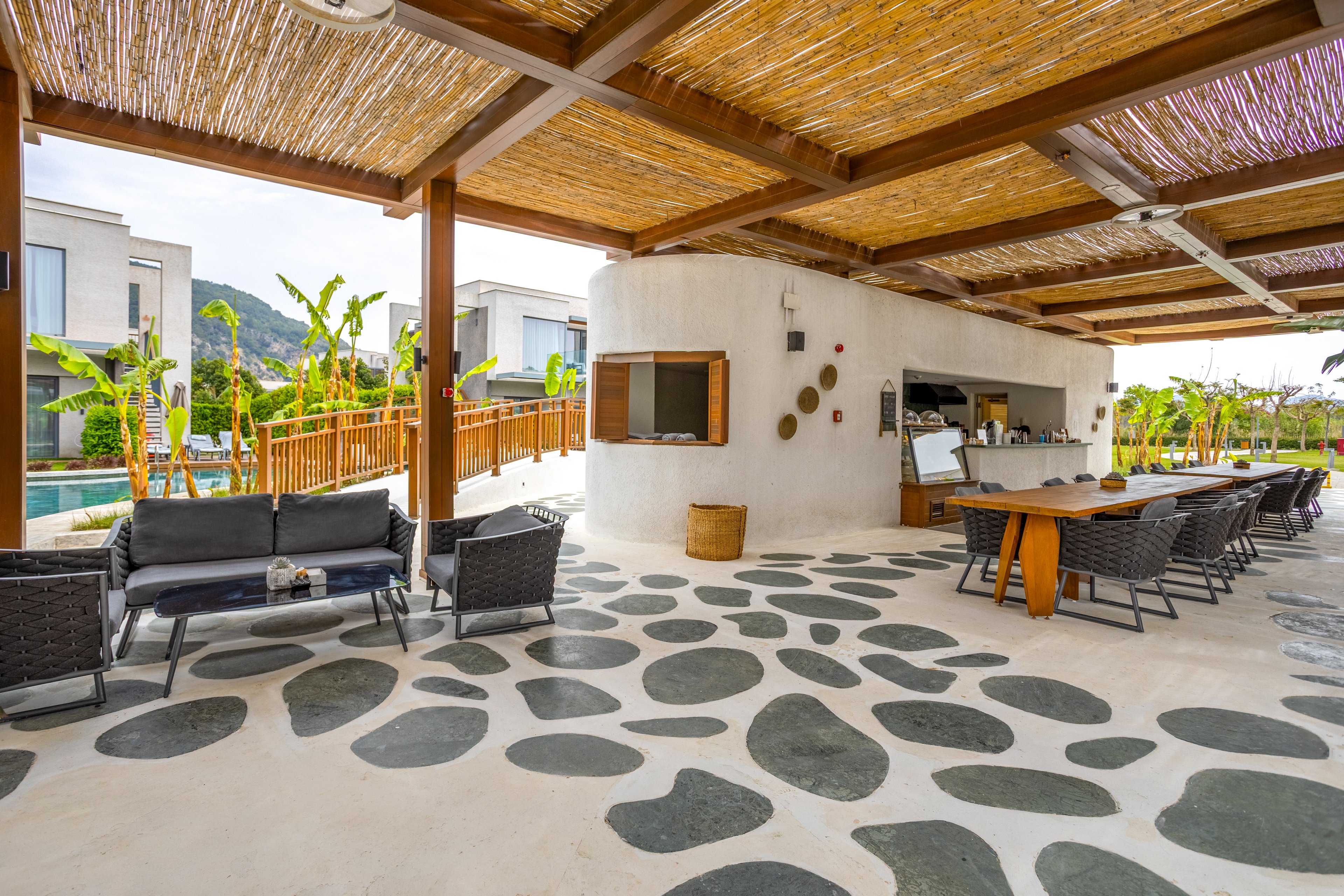 Spacious open-air lounge and dining area features a long table with chairs under a wooden pergola roof. A curved white outdoor kitchen and bar sits at the center, equipped for serving food and drinks. Modern wicker seating and a patterned stone-and-concrete floor create a stylish, relaxed atmosphere. In the background, a pool, tropical plants, and contemporary buildings suggest a resort or vacation property surrounded by greenery and hills.