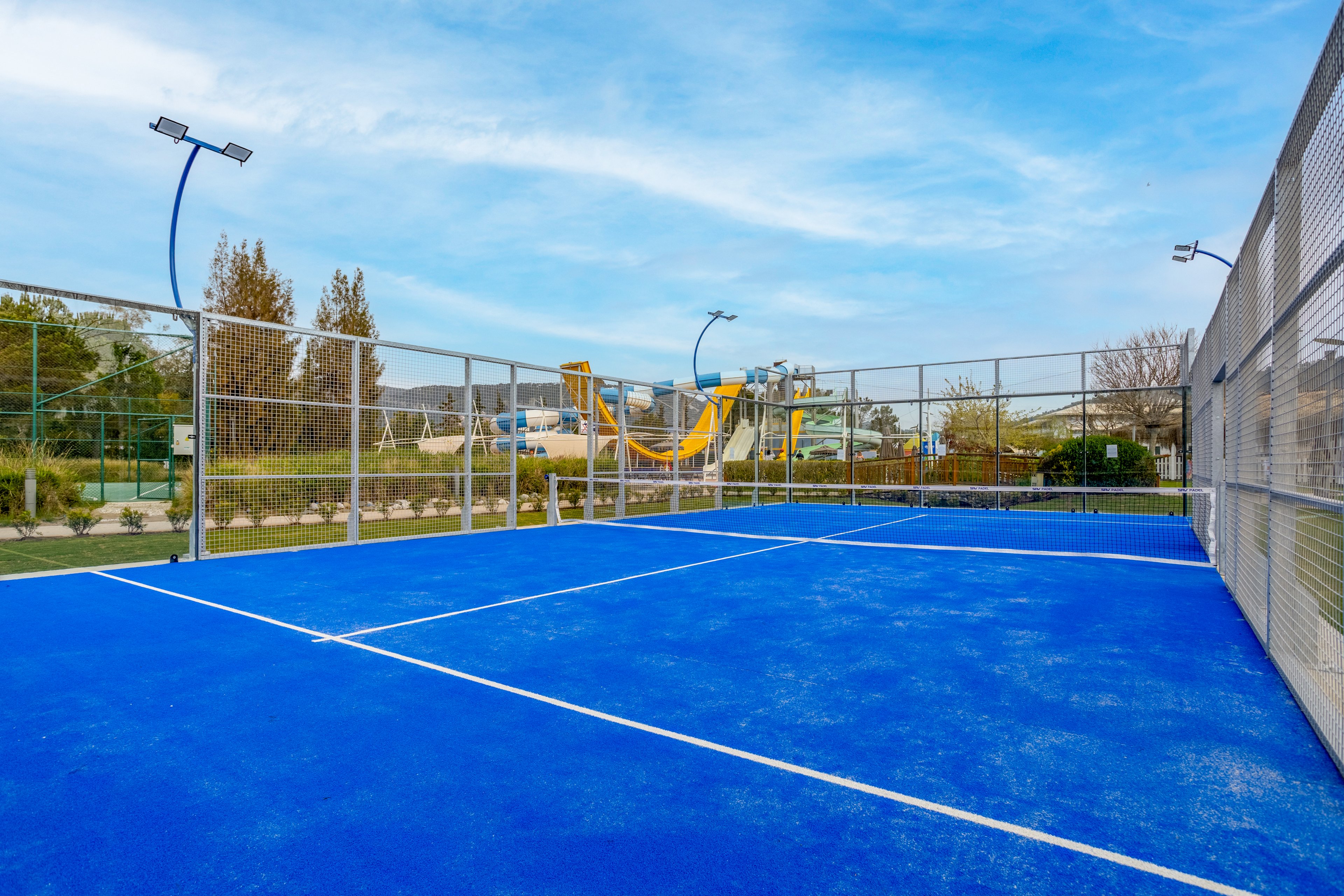 An empty blue tennis court is shown in an outdoor recreational area. The court is surrounded by high metal fencing and modern curved light poles. In the background, there are trees, landscaped greenery, and a colorful playground or slide structure. The sky is clear with soft clouds, giving the scene a bright and inviting atmosphere.