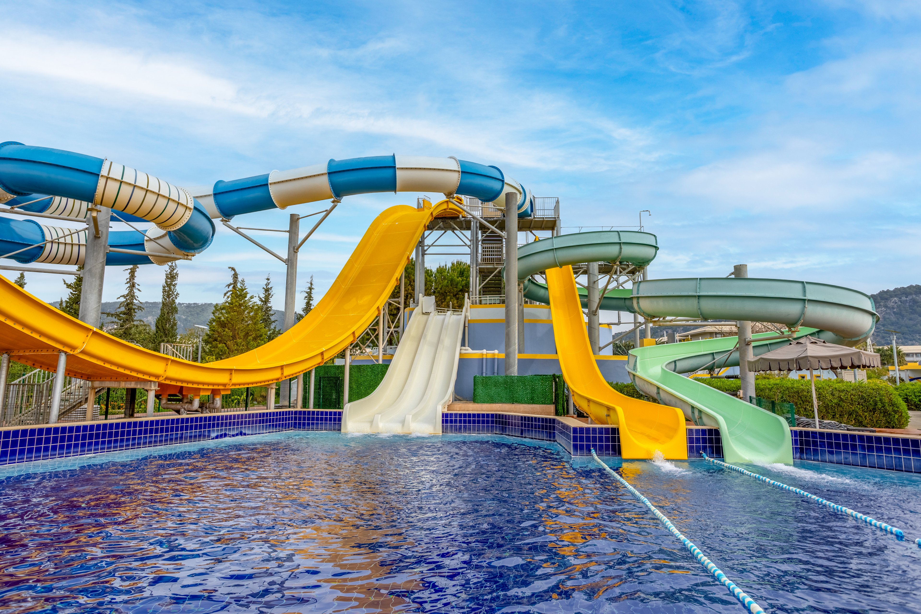 A group of colorful water slides towers above a clear blue swimming pool. The slides include yellow, white, green, and blue striped tubes curving and looping in different directions. The scene is set outdoors with trees, hills, and a partly cloudy blue sky in the background. The image conveys a fun, recreational atmosphere at a water park attraction.