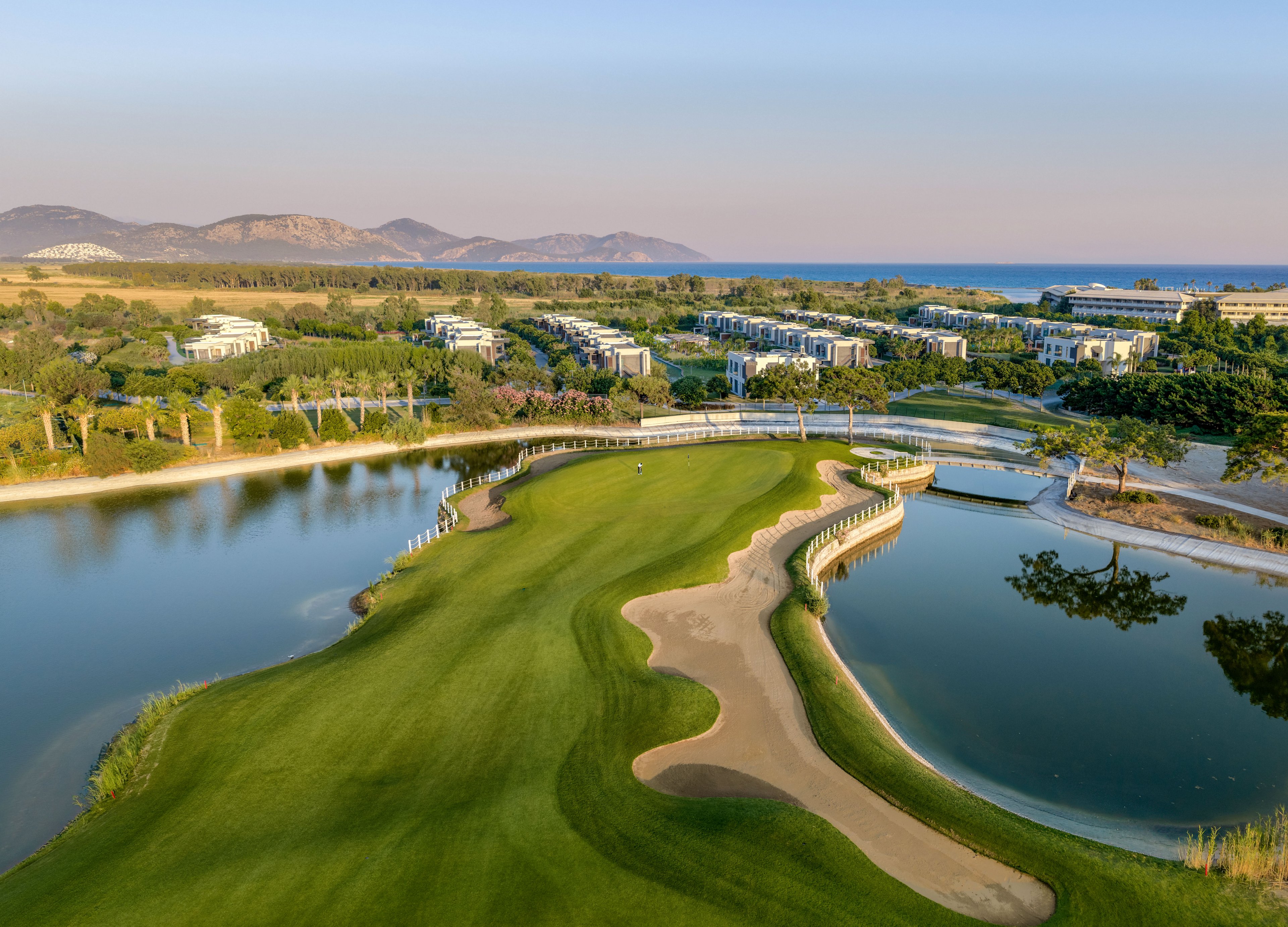 A lush green golf course is surrounded by calm water features and sand bunkers in the foreground. Curved bridges connect the fairway across the ponds, adding visual interest to the landscape. In the distance, modern resort buildings sit among trees with hills and the sea beyond. The scene is captured at golden hour, giving the sky and terrain a warm, tranquil glow.