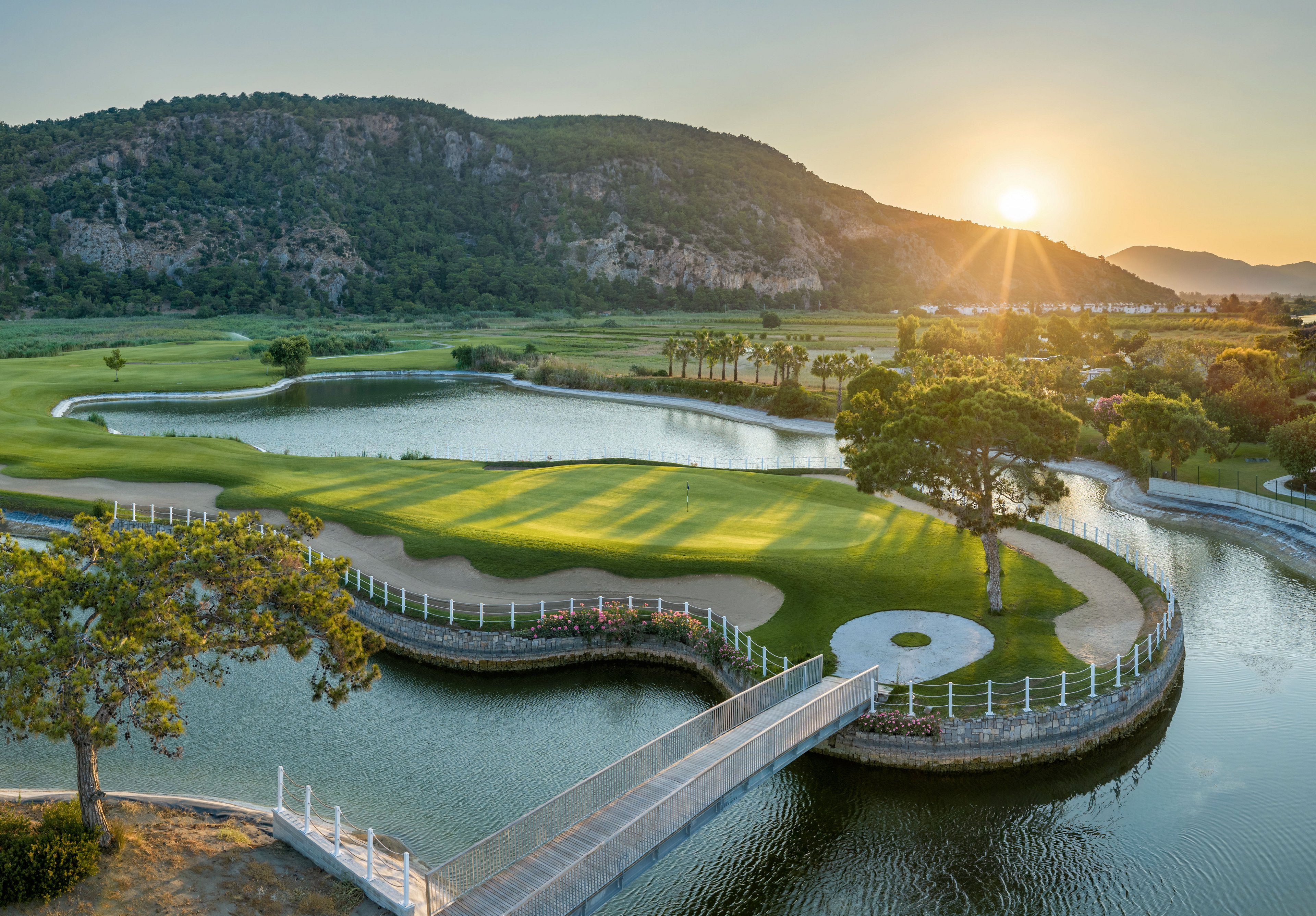 An elevated view shows a lush golf course with an island green surrounded by calm water. A narrow bridge crosses the water, leading to the green bordered by a white fence. Rolling hills and mountains rise in the background under a clear sky. The sun is setting near the horizon, casting warm golden light across the landscape and reflecting on the water.