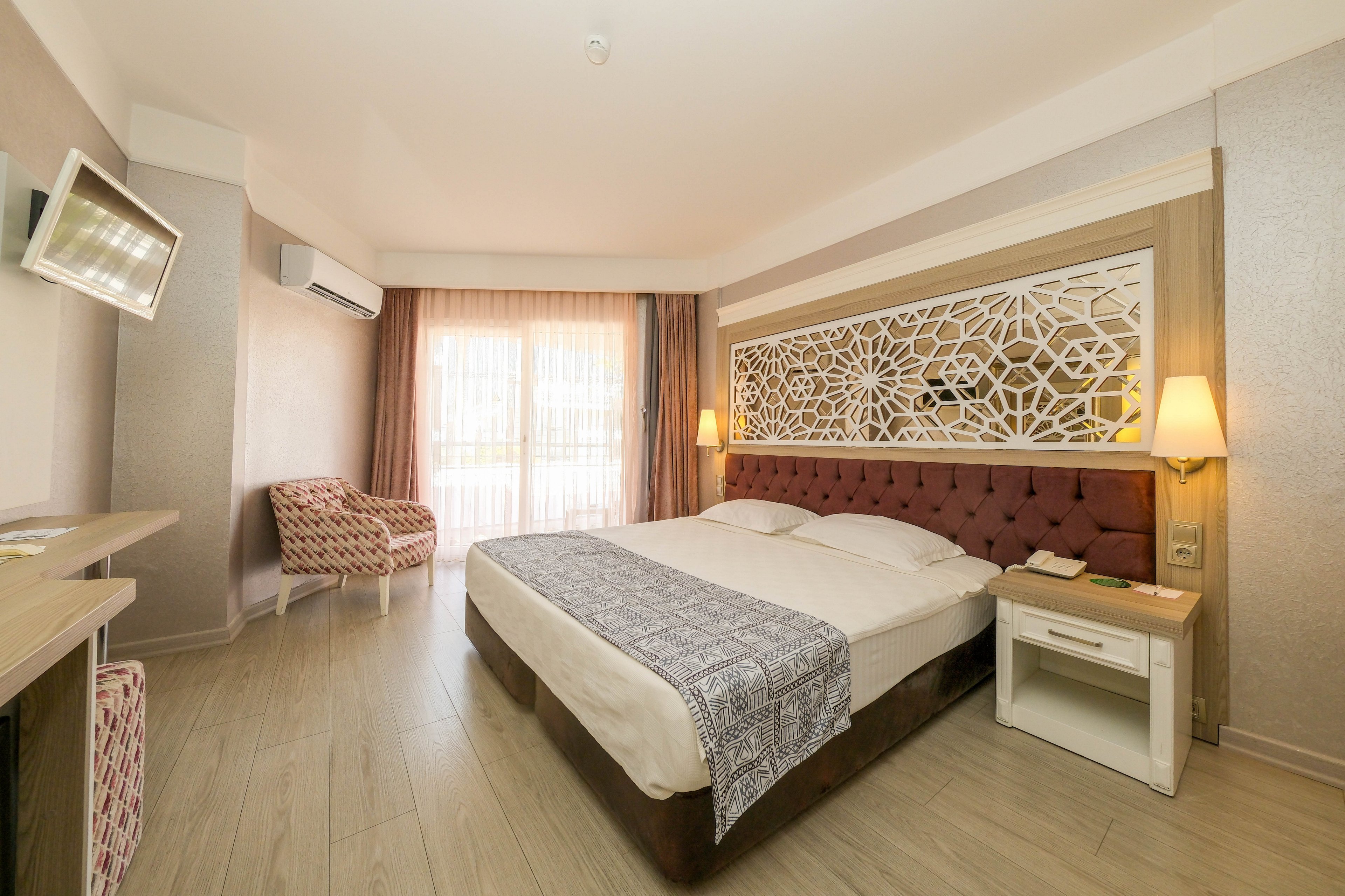 A spacious hotel bedroom features a large double bed with a patterned runner and an ornate geometric headboard. Soft natural light enters through sheer curtains covering a balcony door, illuminating the light wood flooring. A patterned armchair, wall-mounted television, and bedside tables with lamps complete the contemporary decor. The overall atmosphere is clean, warm, and inviting, suitable for comfortable accommodation.