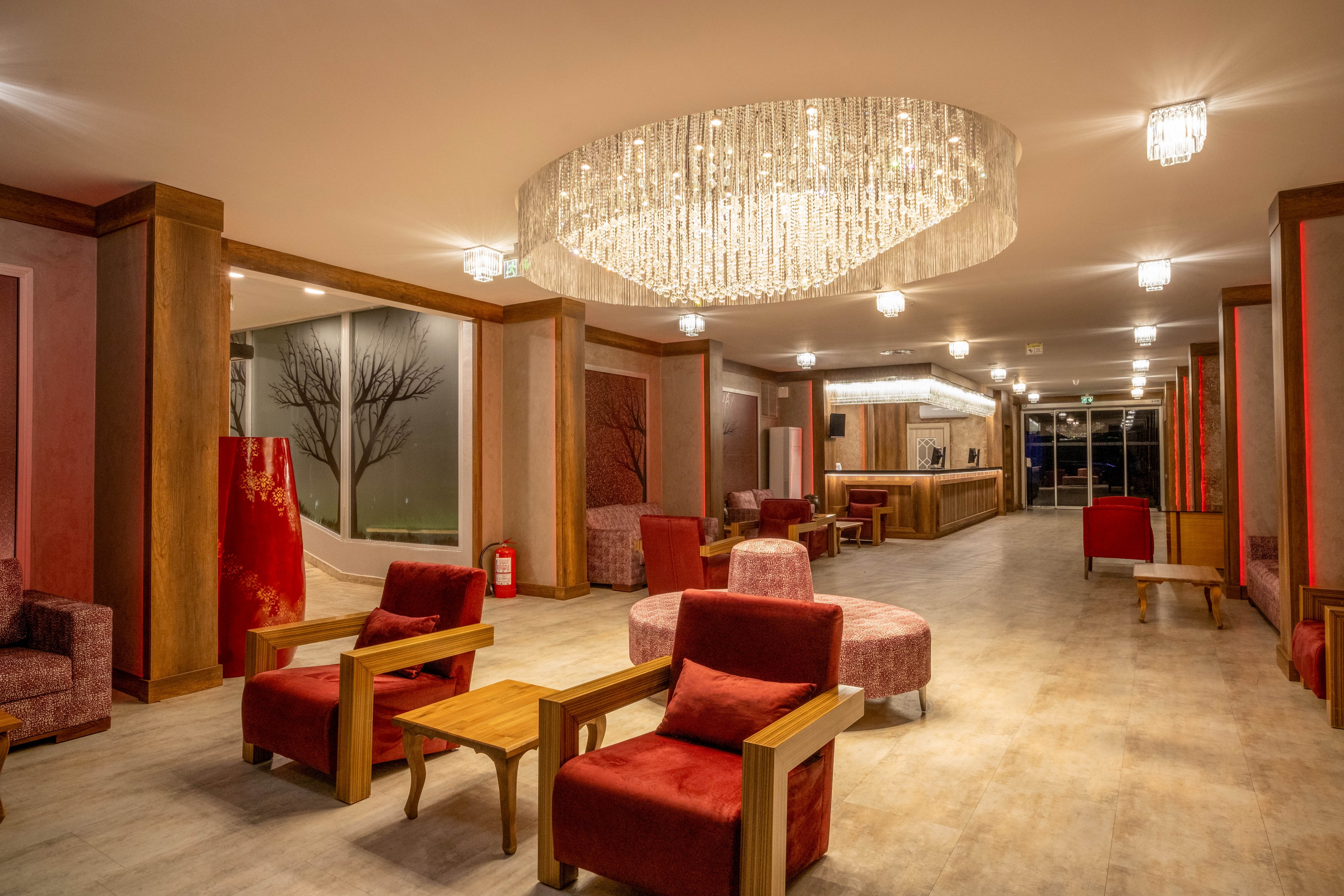 A contemporary hotel lobby features red upholstered armchairs and lounge chairs arranged around wooden tables. Warm lighting from a large crystal chandelier and smaller ceiling fixtures creates a cozy, inviting atmosphere. The space includes a reception desk in the background and decorative wall panels with minimalist artwork. Neutral flooring and wood accents contrast with the bold red tones throughout the interior.