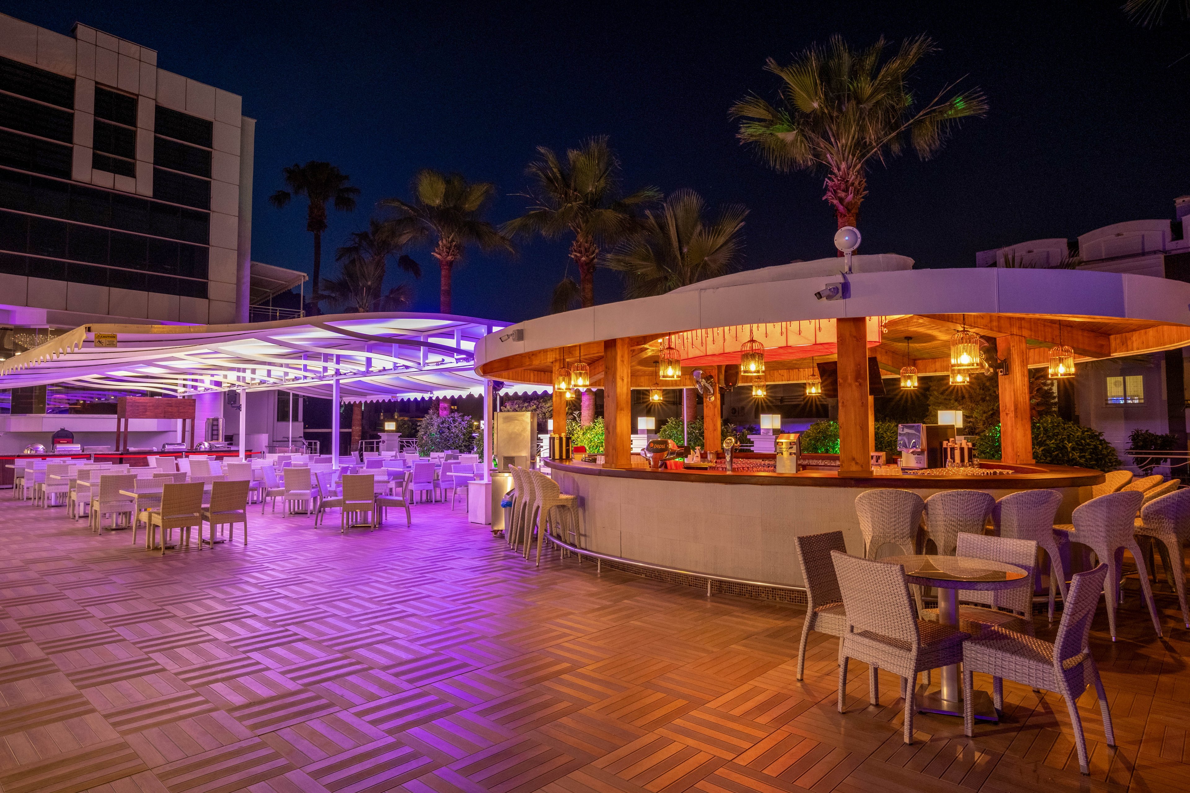 An open-air bar and dining terrace is illuminated with colorful purple and warm yellow lighting at night. The scene features modern wicker chairs and tables arranged on a patterned wooden floor. Palm trees and contemporary buildings frame the space, creating a relaxed vacation atmosphere. The bar counter is highlighted with decorative lights and hanging elements, suggesting a stylish resort or hotel setting.
