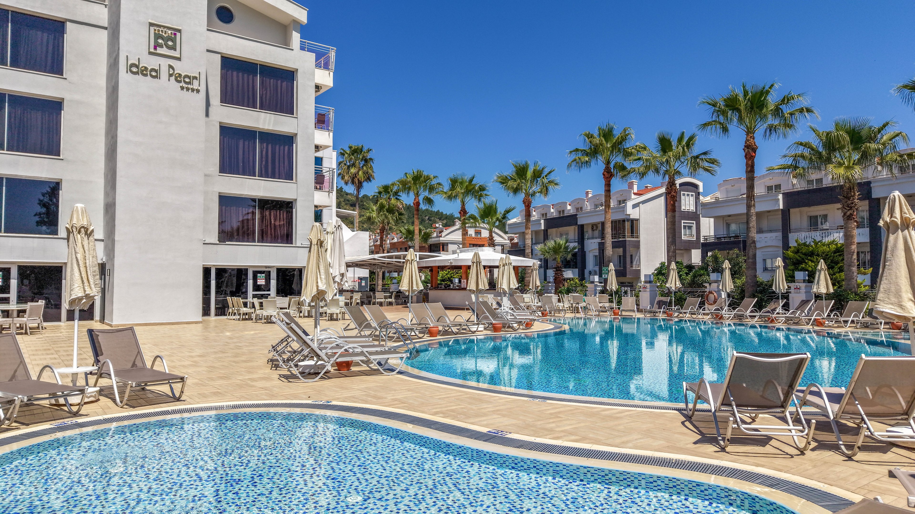 A modern hotel building named Ideal Pearl overlooks a large outdoor swimming pool. Sun loungers and closed beige umbrellas surround the pool on a tiled terrace, suggesting a relaxing vacation setting. A smaller mosaic-tiled pool is visible in the foreground, with palm trees and neighboring buildings in the background under a clear blue sky. The scene conveys a bright, sunny, resort atmosphere with ample seating and leisure space.
