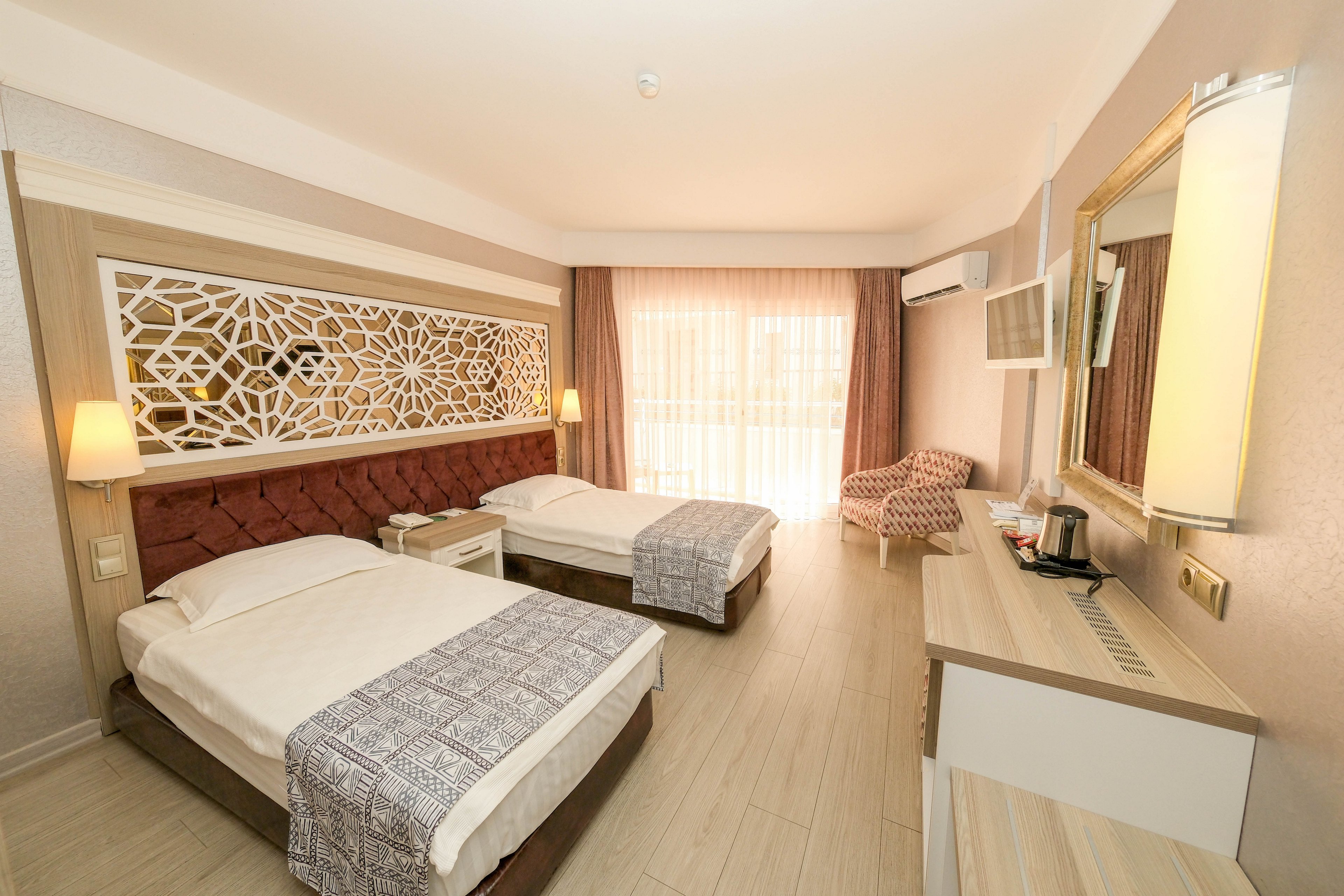A contemporary hotel room features two neatly made twin beds with patterned runners and a decorative lattice headboard. The space includes bedside tables with lamps, a desk with a kettle and cups, and a cushioned armchair near the wall. Large curtains cover a glass door or window, allowing soft natural light to fill the room. Neutral tones and clean lines create a calm, inviting atmosphere suitable for guests.