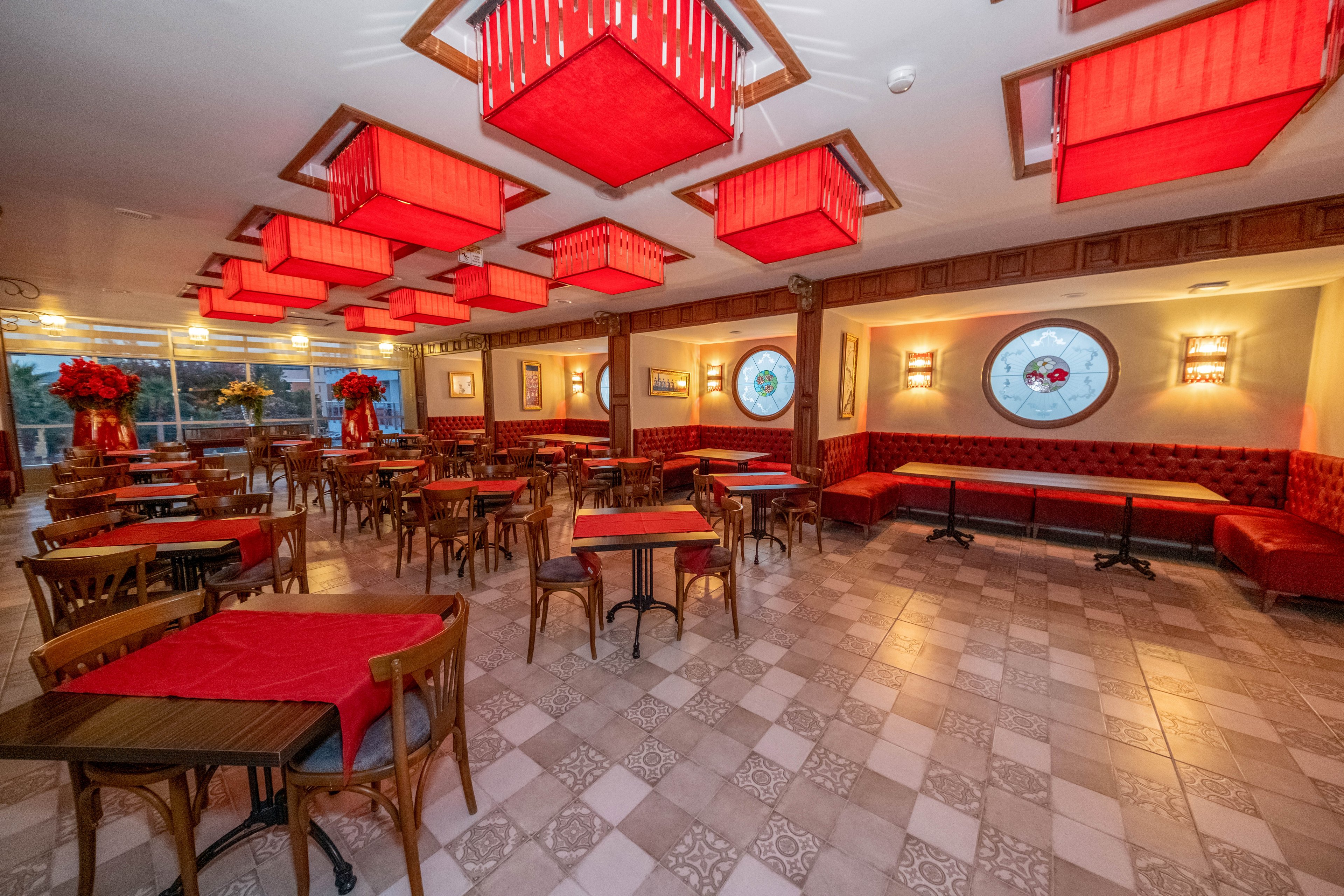 The image shows an empty restaurant dining area with wooden tables and chairs neatly arranged. Red tablecloths, upholstered bench seating, and large red ceiling light fixtures create a warm, vibrant atmosphere. Decorative circular stained-glass windows and wall sconces add visual interest along the side wall. The tiled floor features a subtle patterned design, and large windows at the back let in natural light.