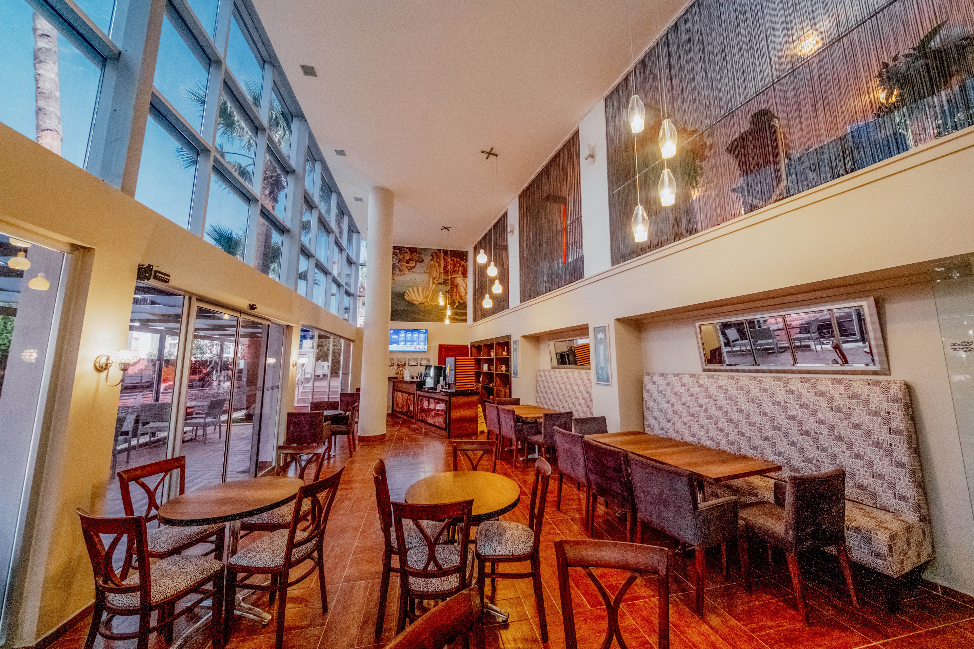 The image shows a contemporary cafe or restaurant interior with wooden tables and upholstered chairs arranged on a polished wood floor. Large floor-to-ceiling glass windows on the left side let in natural light and reveal an outdoor seating area. Pendant lights hang from the high ceiling, creating a warm and inviting atmosphere. Artwork and decorative wall panels add visual interest to the upper level, while a counter or bar area is visible toward the back of the space.