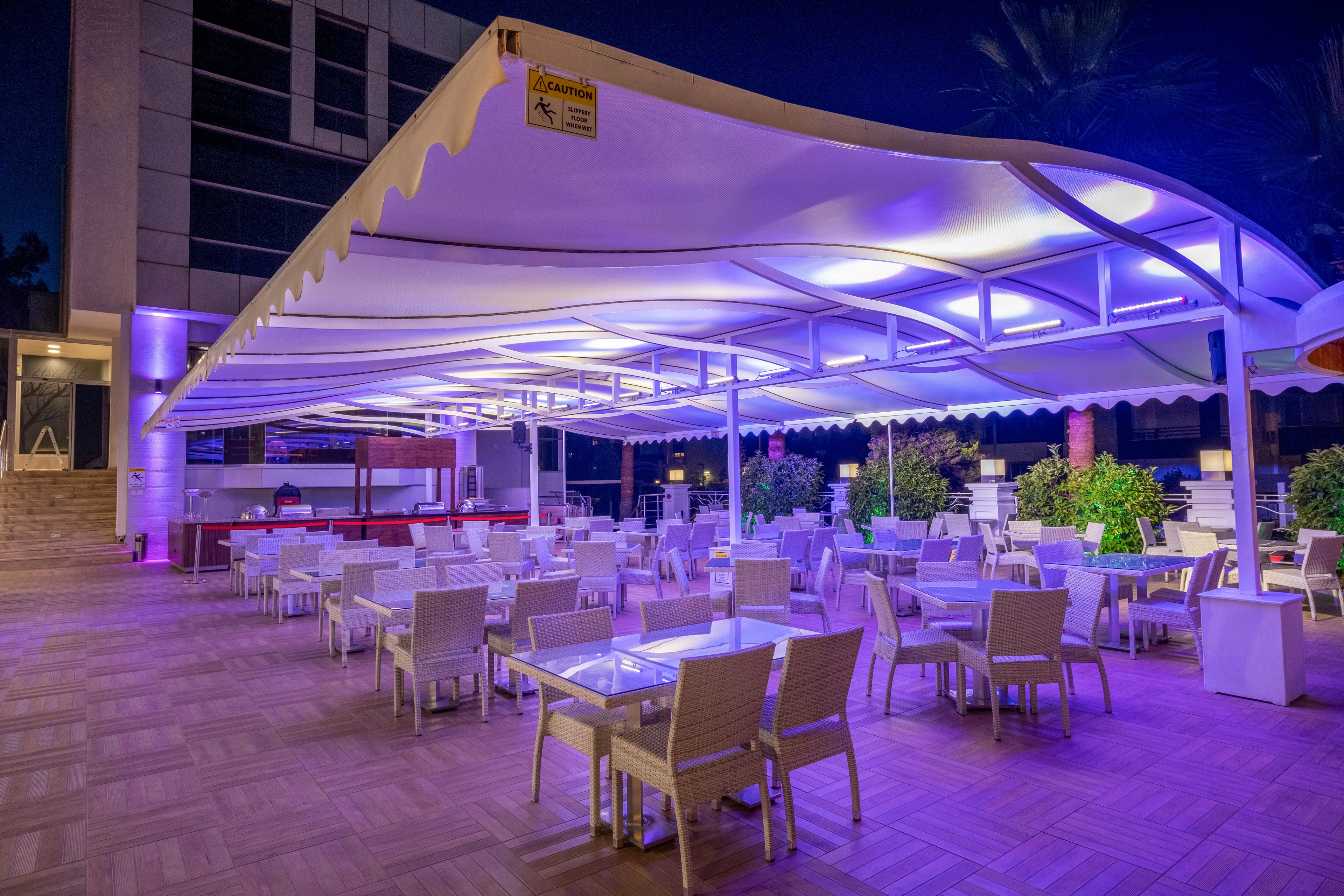 An open-air restaurant terrace is shown at night with rows of white tables and chairs neatly arranged under a large canopy. The scene is illuminated by vibrant purple and blue lights, creating a modern and inviting atmosphere. Potted plants line the edges of the seating area, and a bar or service counter is visible in the background. The surrounding building and palm trees suggest a contemporary hotel or resort setting.