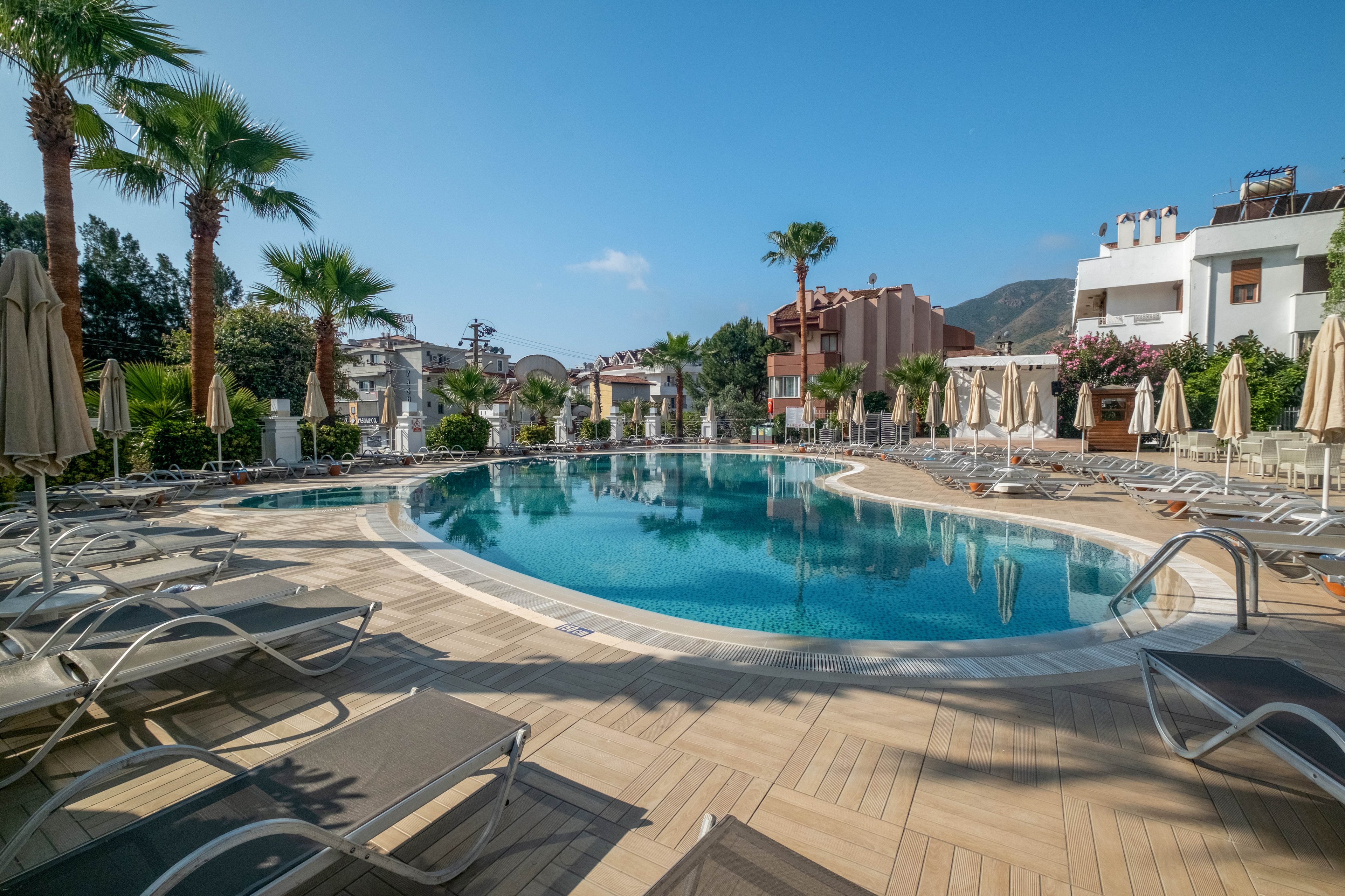 A curved outdoor swimming pool is surrounded by numerous sun loungers and beige umbrellas at a resort. Palm trees and flowering shrubs frame the pool area, adding a tropical feel. Modern multi-story buildings and distant hills are visible in the background under a clear blue sky. The scene suggests a quiet, relaxing vacation setting in warm weather.