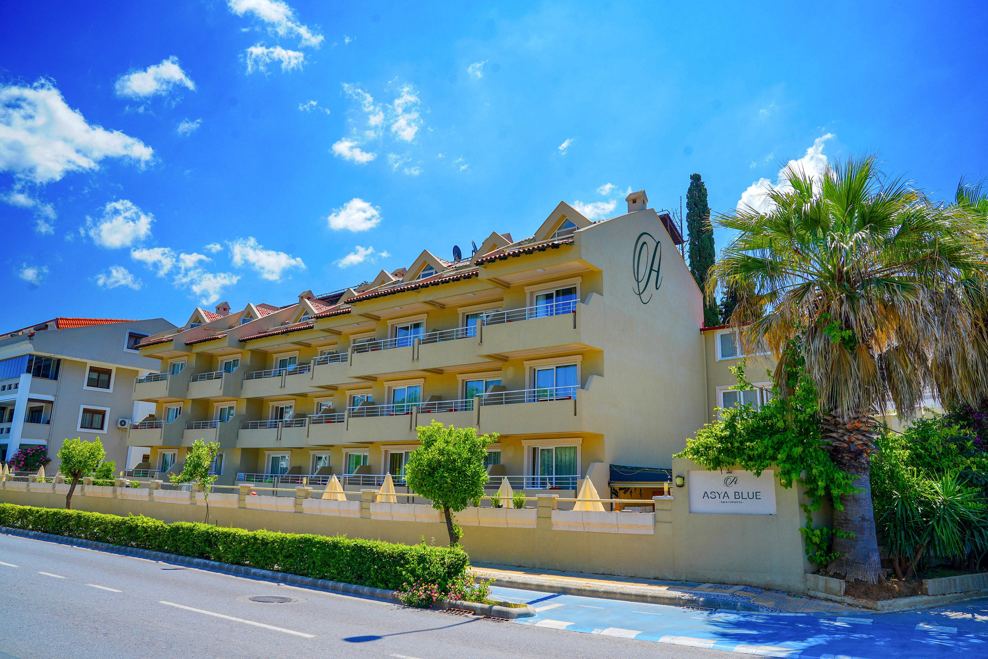 A multi-story hotel building with rows of balconies stands along a quiet street under a bright blue sky. Palm trees and neatly trimmed shrubs line the front of the property, adding a tropical feel. The light-colored facade contrasts with the vivid blue of the sky and bike lane. A sign near the entrance suggests the property name, while the overall scene conveys a relaxed vacation atmosphere.