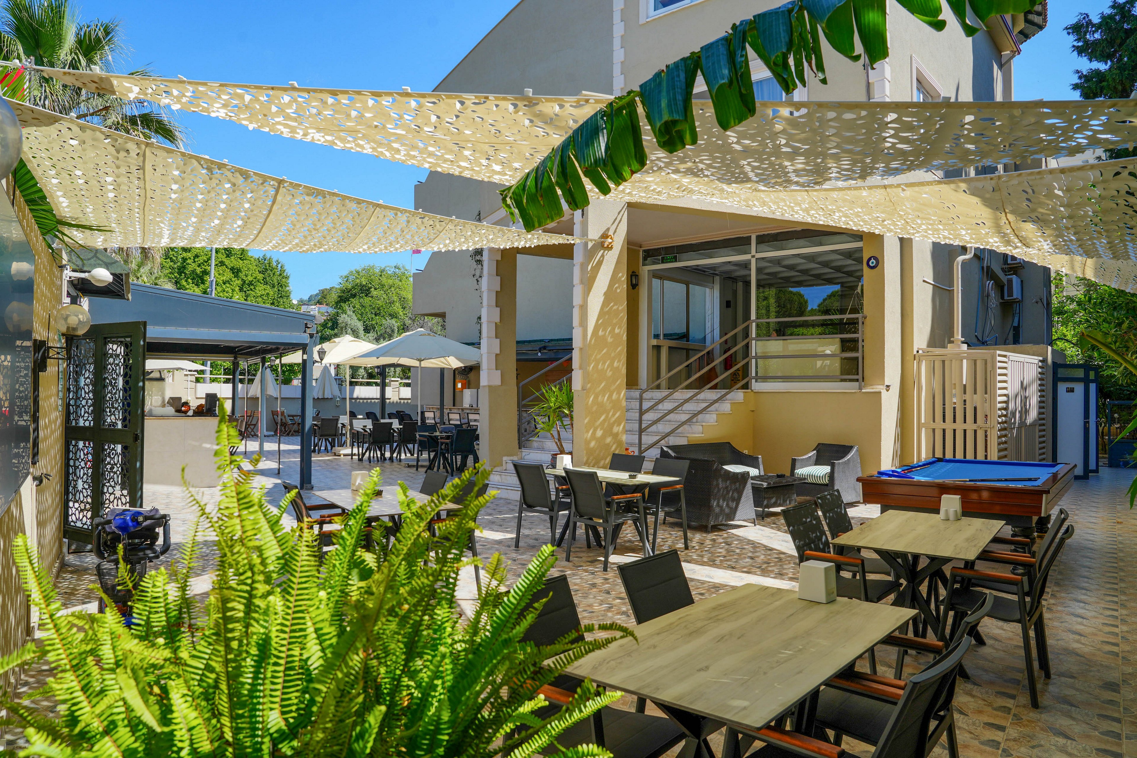 An inviting outdoor cafe terrace features several tables and chairs arranged on a tiled patio. Light fabric shade sails are stretched overhead, casting patterned shadows on the seating area. Lush green plants and trees surround the space, adding a fresh, natural atmosphere. In the background, additional covered seating and a modern building facade complete the relaxed, open-air setting.