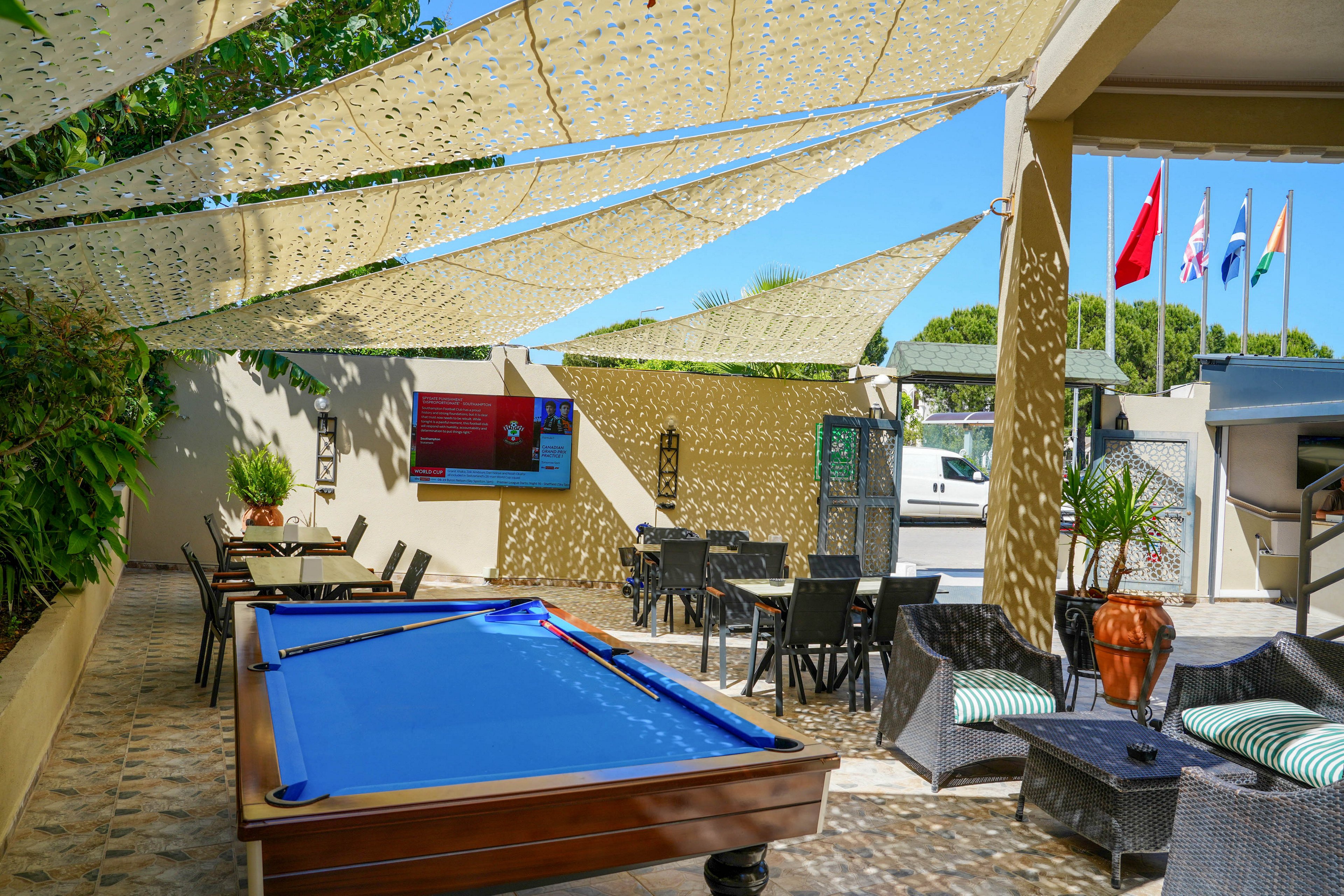 An outdoor leisure area features a bright blue pool table as the main focal point. Shaded canopies stretch overhead, casting patterned shadows across the tiled floor and seating area. Wicker chairs and tables create a casual lounge atmosphere beside potted plants and greenery. In the background, several national flags fly near a parking area under a clear blue sky.