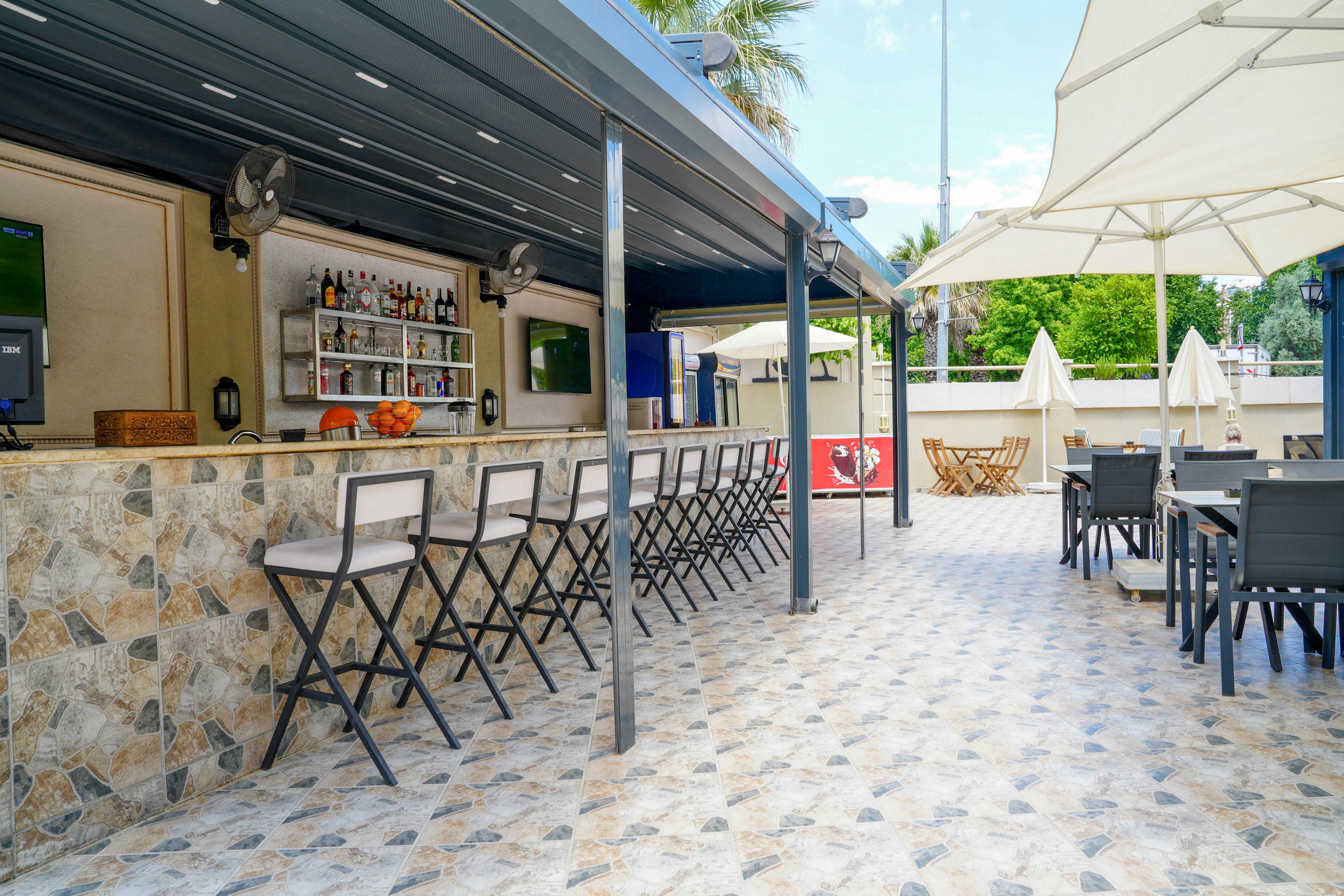 An open-air bar with a stone counter and high stools stands ready for guests in a sunny outdoor setting. Bottles and glasses are neatly arranged on shelves behind the bar, with fruit displayed on the counter. Modern tables and chairs are set up under large white umbrellas on a patterned tiled floor. The scene is bright and inviting, suggesting a relaxed vacation or resort atmosphere.