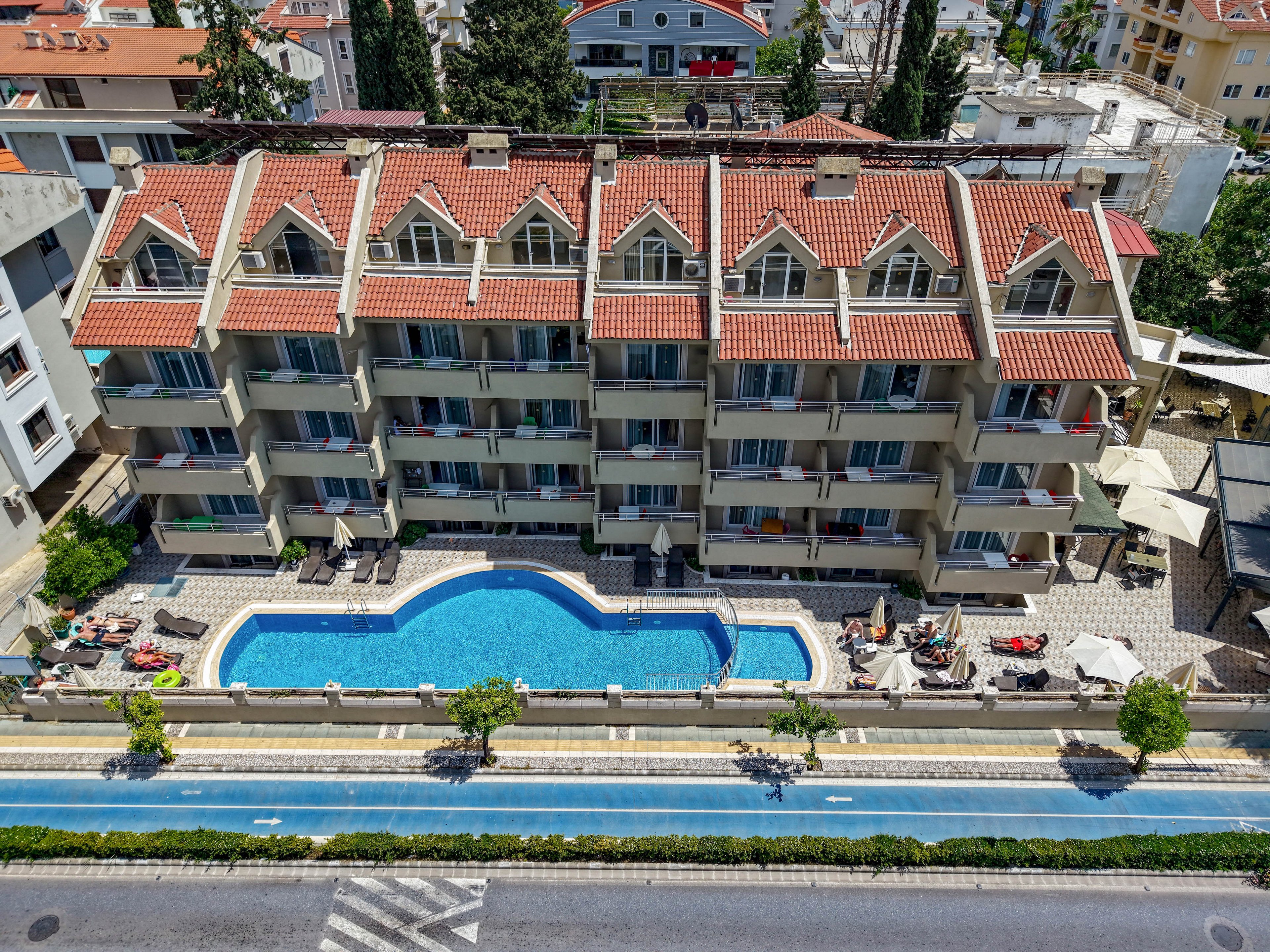 A multi-story apartment or hotel complex with red-tiled roofs surrounds a central outdoor swimming pool. The scene is captured from above, showing balconies facing the pool and several people relaxing on sun loungers. A narrow decorative water feature or secondary pool runs parallel to a road in the foreground. The surrounding area includes neighboring buildings, trees, and a bright, sunny atmosphere.
