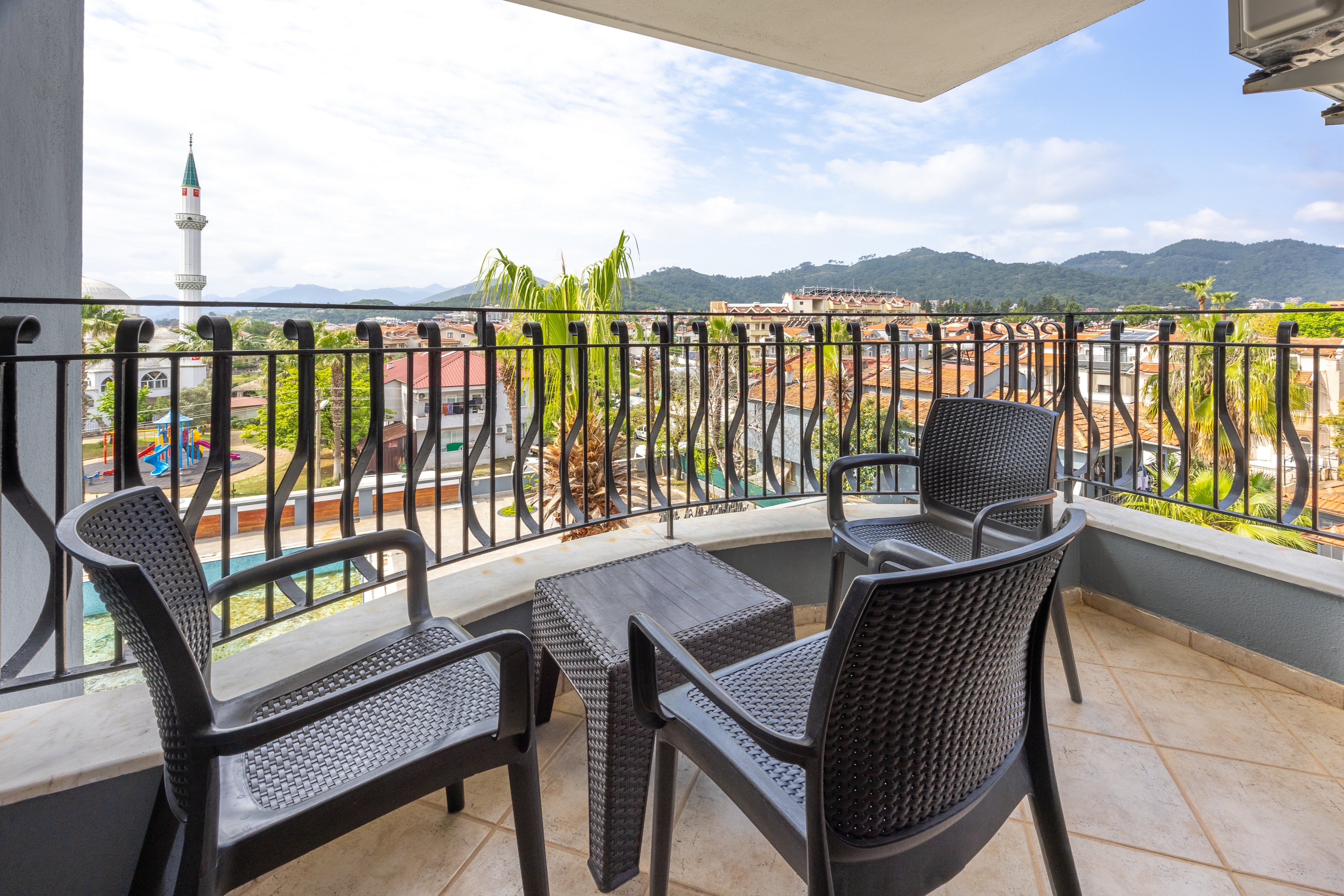 A modern balcony features three black chairs and a small table, overlooking a scenic cityscape with mountains in the background. The setting is bright and airy, with tiled flooring and decorative railing. A mosque minaret is visible in the distance, adding a cultural touch to the urban landscape. The overall mood is relaxed and inviting, ideal for leisure or conversation.
