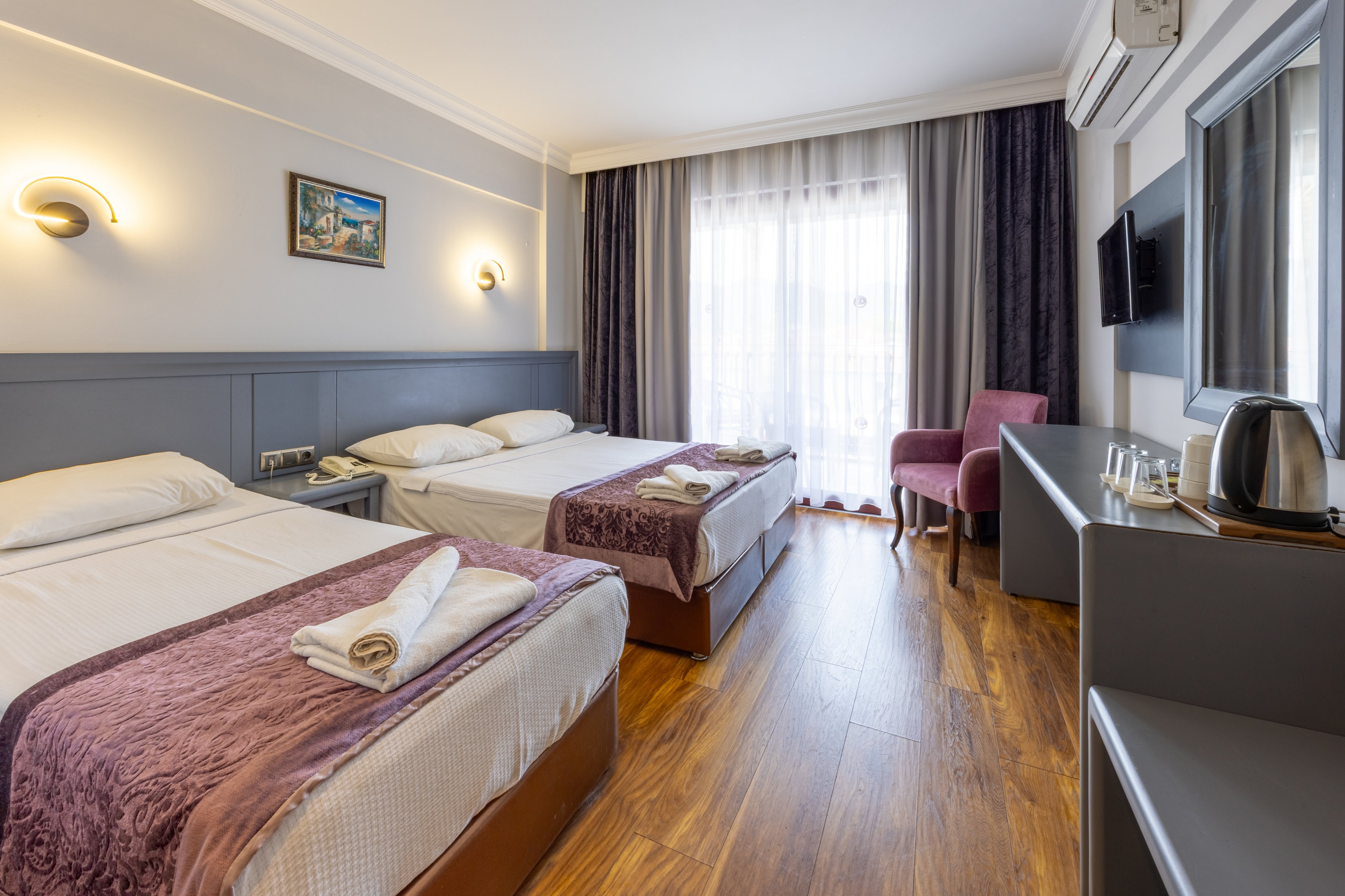 A contemporary hotel room features two neatly made beds with folded towels, set against a soft gray and lavender palette. The space includes a large window with sheer curtains, a small balcony, and a cozy seating area with a mauve chair. Amenities such as a wall-mounted TV, electric kettle, and complimentary water are visible on the side console. The room is well-lit with natural light and modern wall sconces, creating a welcoming and comfortable atmosphere.