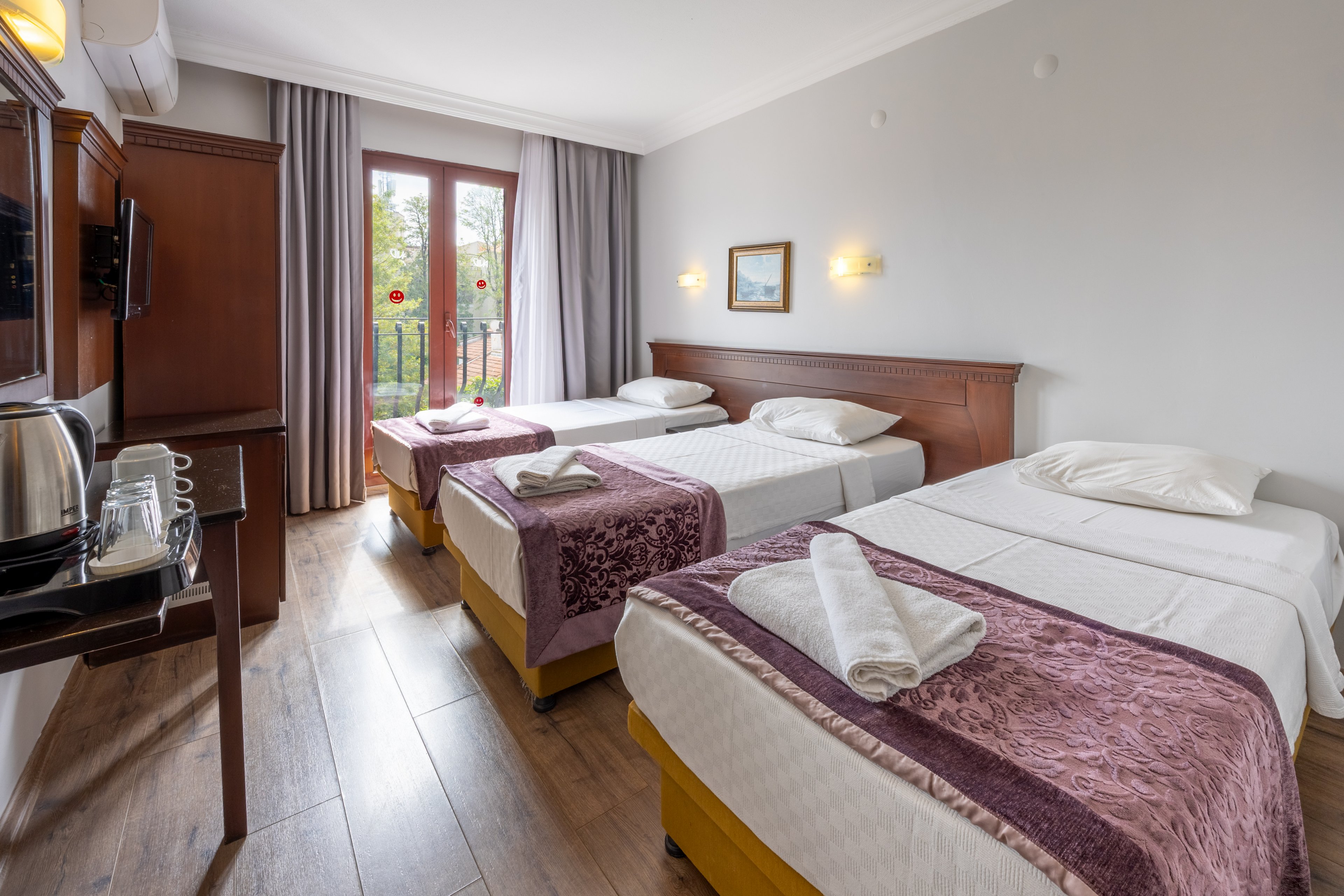 A spacious hotel room features three single beds neatly arranged with folded towels and purple throws. The room is well-lit with natural light from a glass door leading to a balcony overlooking greenery. Modern amenities include a wall-mounted TV, kettle set, and wooden furnishings. The decor is simple with neutral walls, framed art, and wood flooring. No visible text or numbers are present in the image.