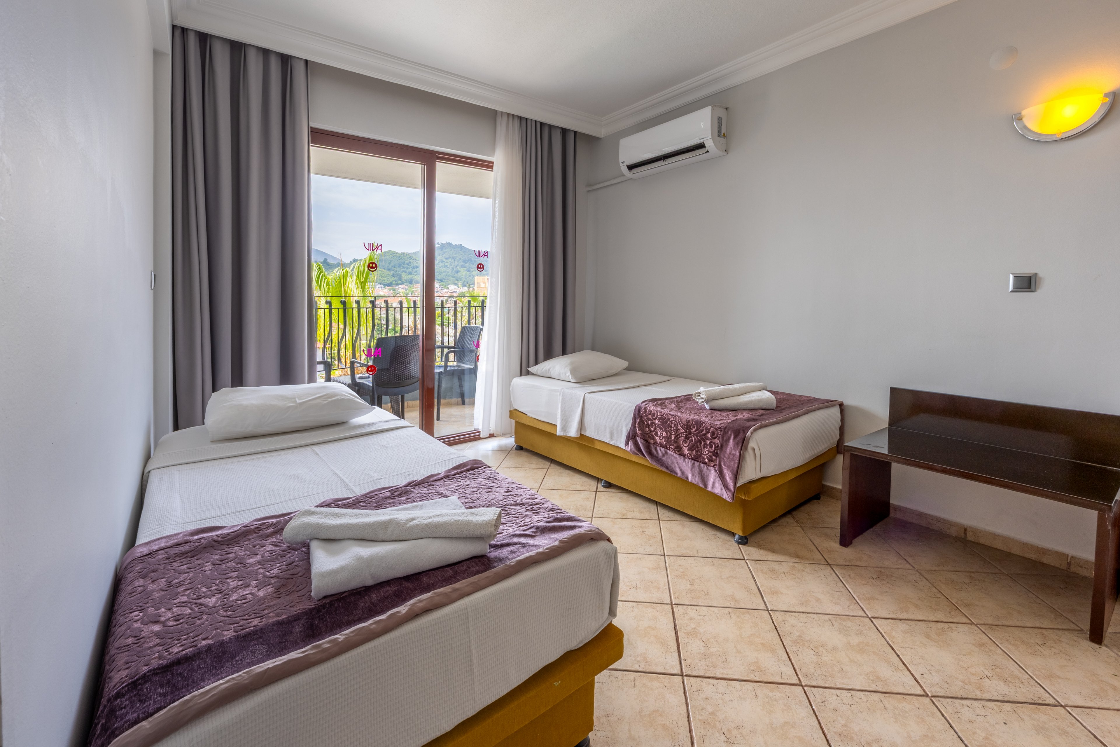 A modern hotel room features two single beds with white linens and folded towels, accented by light purple throws. The space is well-lit with natural light from a sliding glass door that opens to a balcony overlooking greenery and distant hills. The room includes tiled flooring, a wall-mounted air conditioner, and a small wooden table. The overall palette is neutral with soft purple and yellow tones, creating a clean and inviting atmosphere. No visible text or numbers are present in the image.