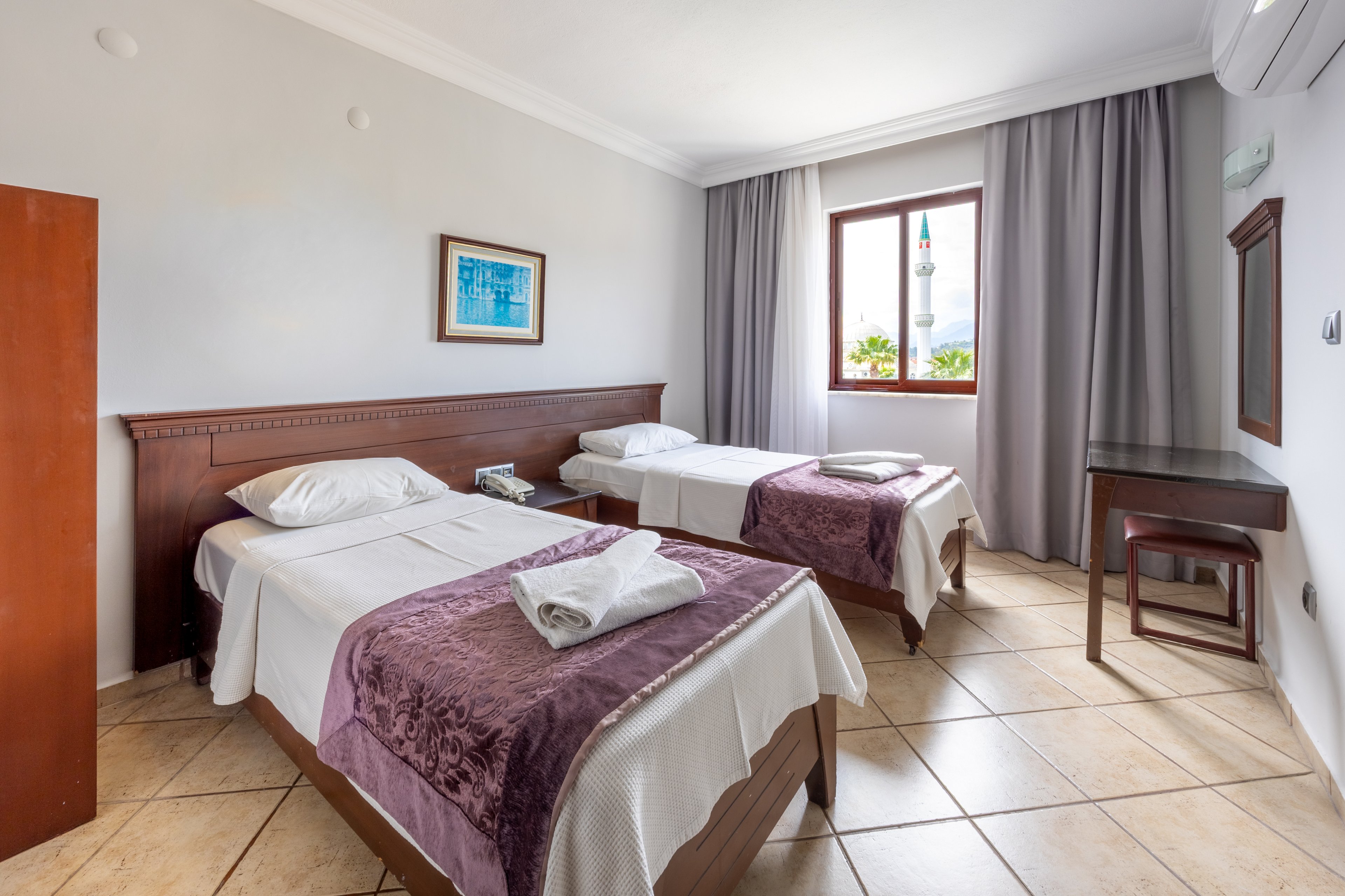 A clean, well-lit hotel room features two single beds with white linens and purple throws. The room includes a wooden headboard, a small desk with a mirror, and a window with sheer and blackout curtains letting in natural light. Tiled floors and minimal decor create a fresh, uncluttered atmosphere. A framed artwork hangs above the beds, and folded towels are placed on each bed. No visible text or numbers are present in the image.