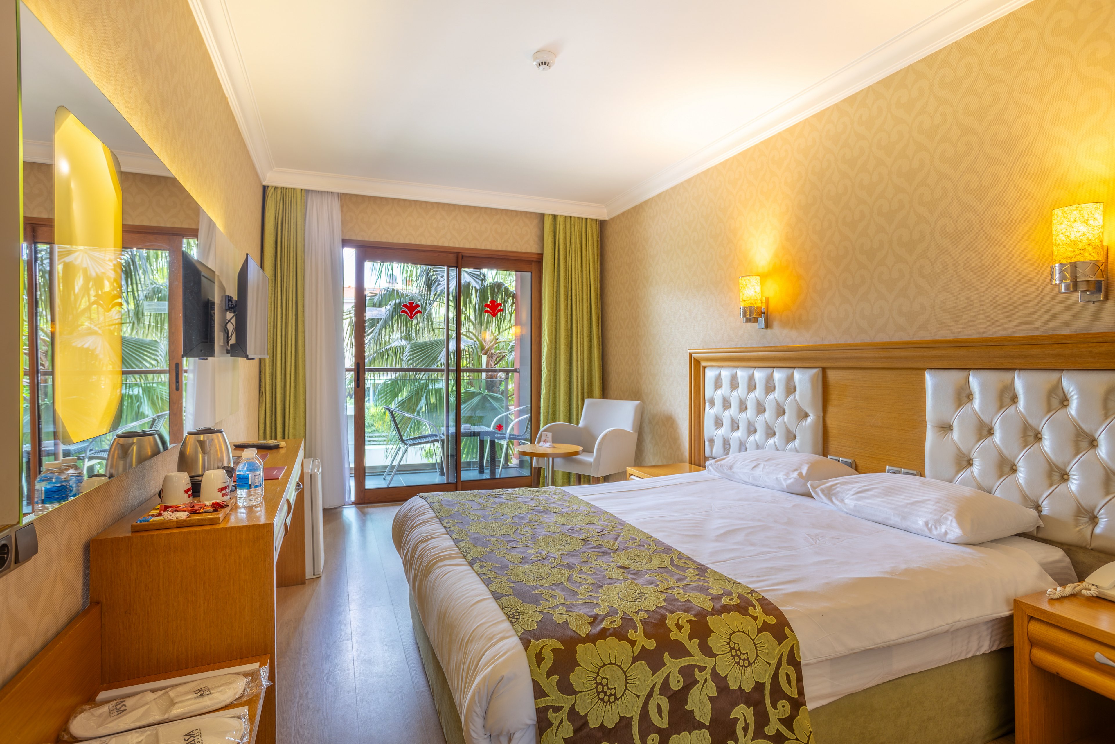 A neatly made double bed dominates a bright hotel room with upholstered headboard and patterned runner. Warm yellow walls and soft lighting from wall sconces create a cozy, inviting atmosphere. A desk with a flat-screen TV, bottled water, and amenities lines one side of the room. Glass doors open to a balcony with chairs, overlooking lush palm trees and greenery outside.