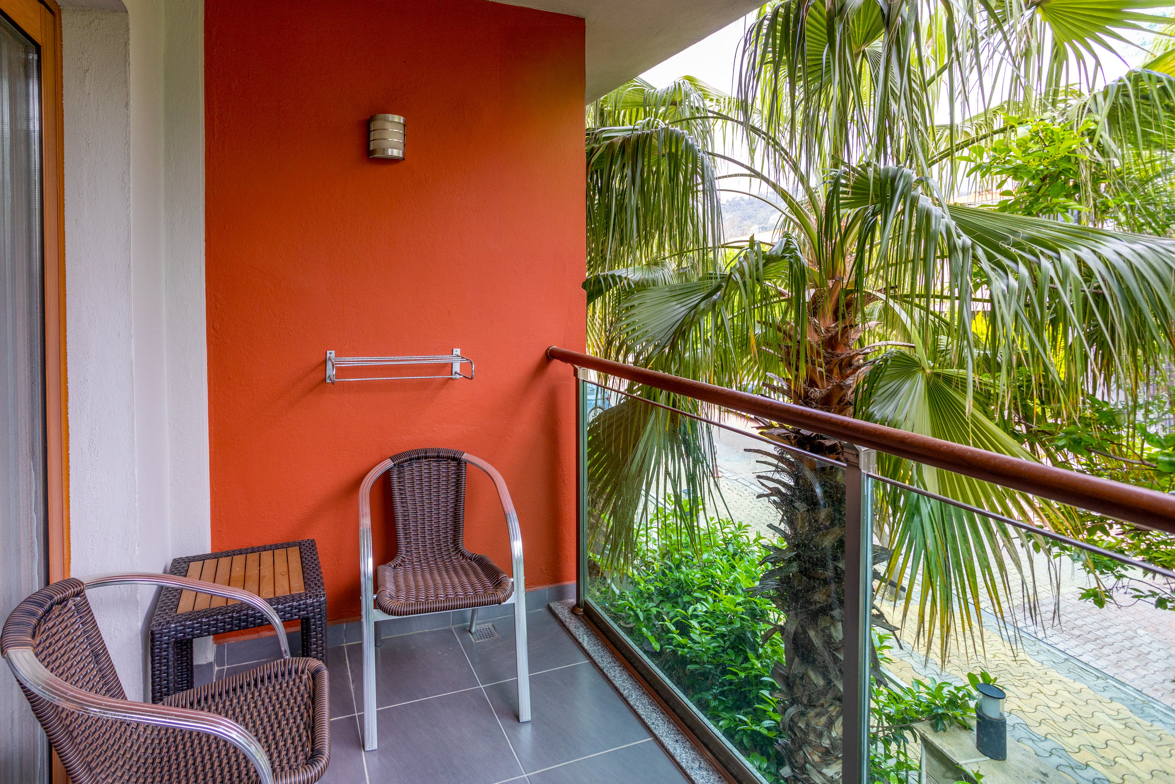 A small balcony features two brown wicker chairs and a matching side table against an orange wall. The space overlooks lush green palm trees and a paved walkway below. Glass railing panels with metal supports frame the balcony, allowing an open view of the surrounding greenery. The scene suggests a quiet outdoor spot for relaxation at a hotel or apartment.