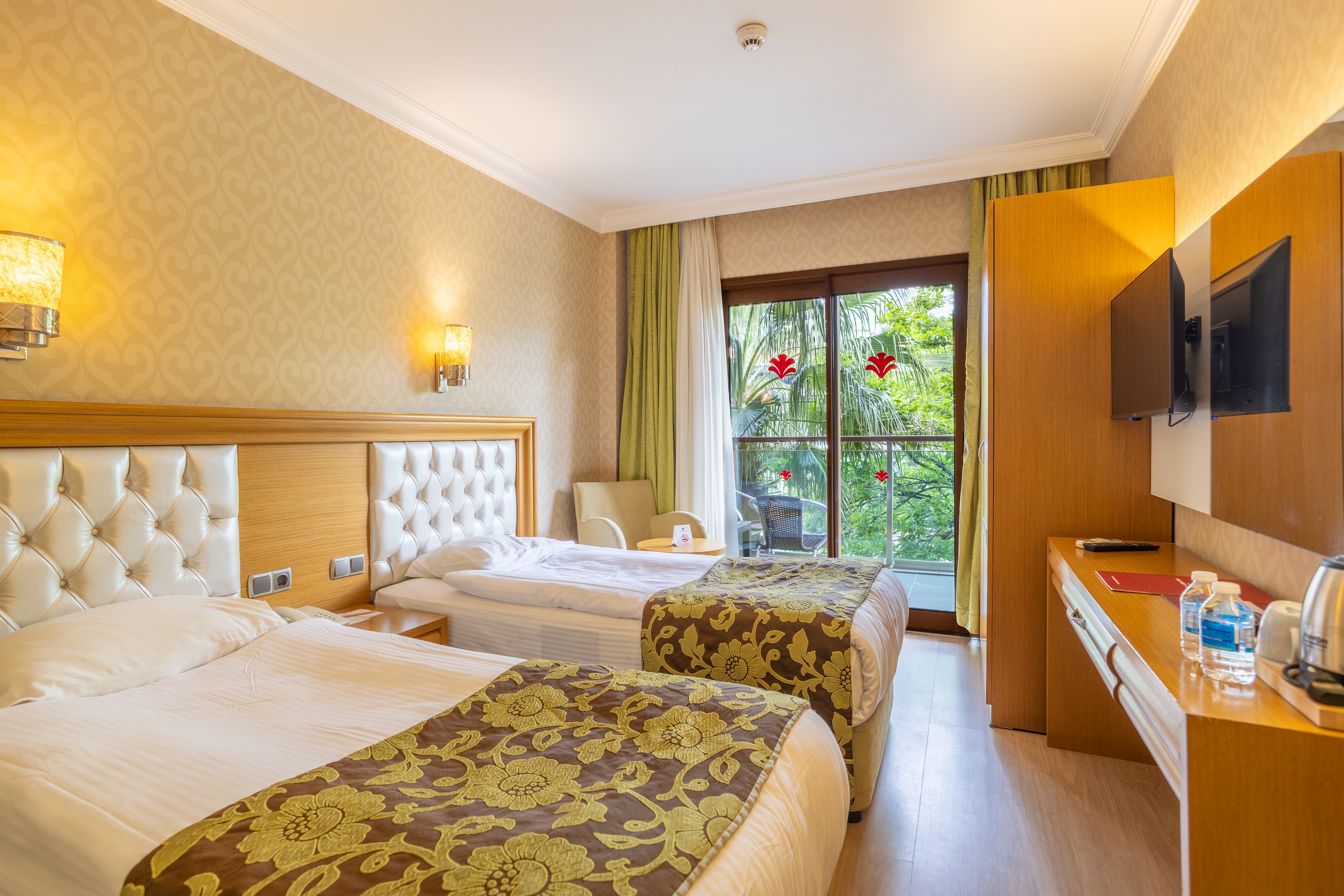 A cozy hotel room features two single beds with tufted headboards and patterned bed runners. Warm wall sconces and neutral wallpaper create a welcoming atmosphere. A desk area with a flat-screen TV, bottled water, and a chair sits opposite the beds. Large glass doors open to a balcony with a view of lush green palm trees and hanging red decorations.