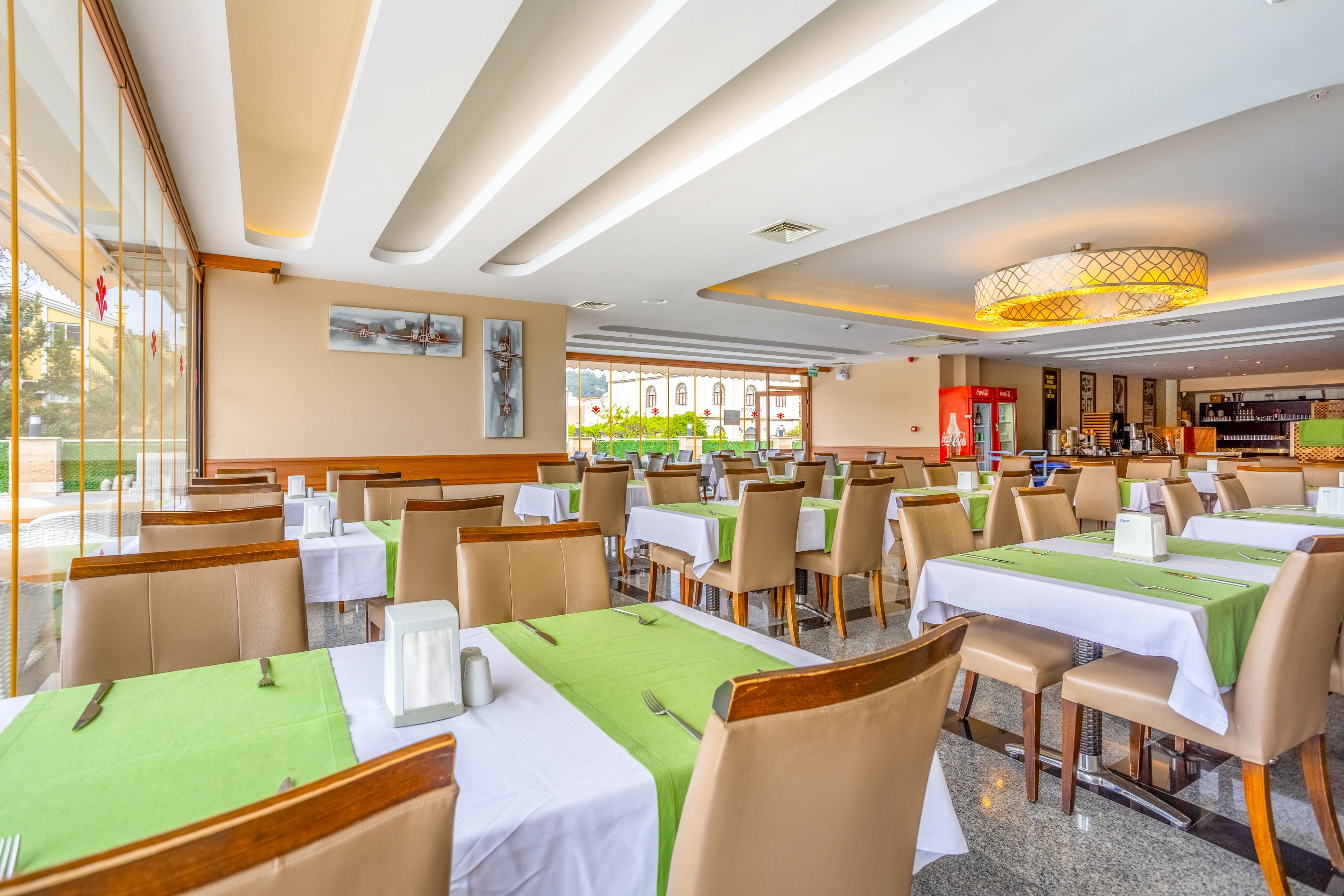 A spacious modern restaurant dining area features neatly arranged tables covered with white cloths and bright green runners. Beige upholstered chairs surround the tables, which are set with napkin holders and condiments. Large windows on one side let in abundant natural light, illuminating the polished floor and contemporary ceiling design. In the background, a bar and beverage area add depth to the open, inviting space.