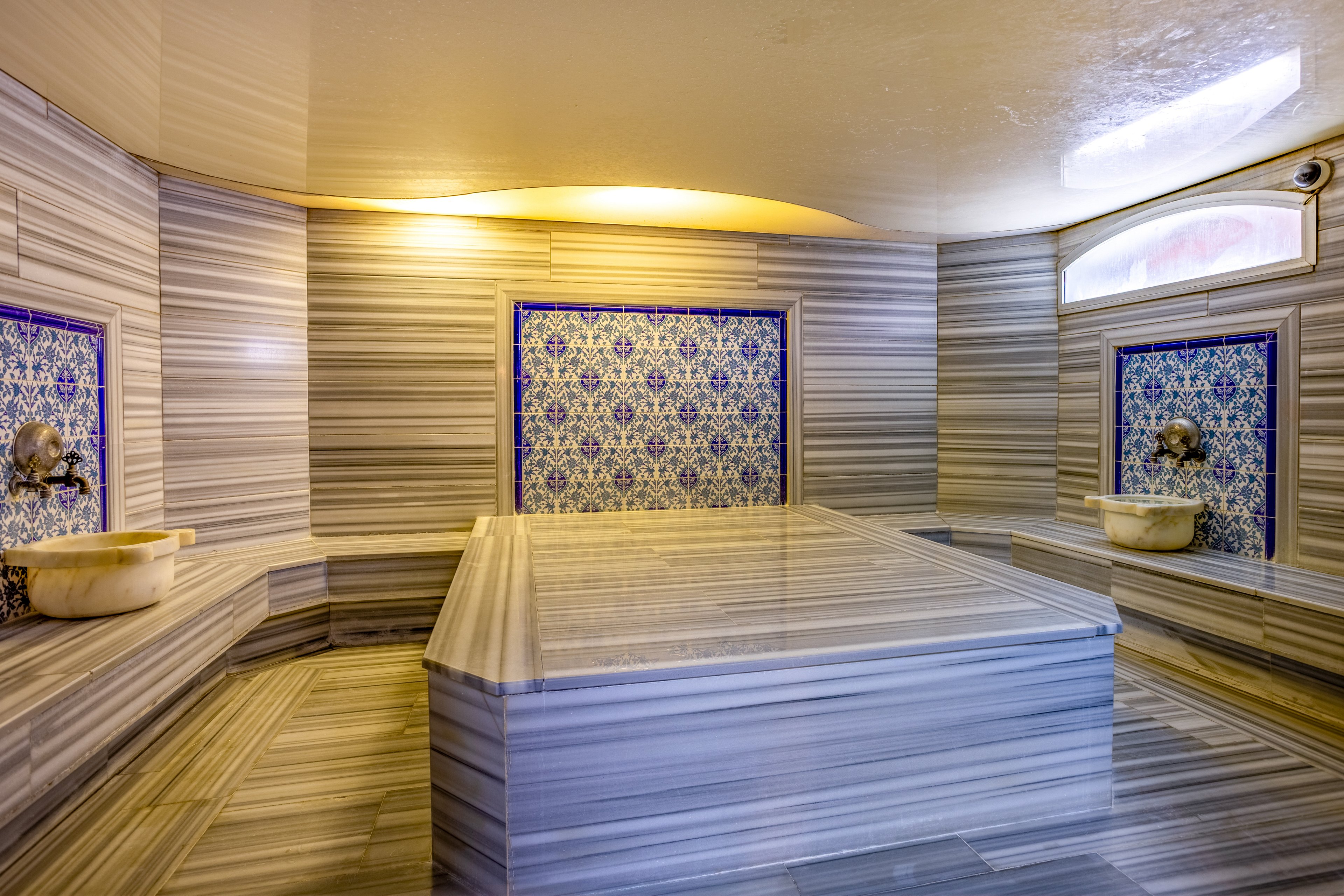 Luxurious Turkish hammam interior featuring a large central marble platform. The room is lined with striped gray marble walls and floors, creating a clean, polished look. Ornate blue and white tiled panels with decorative faucets frame marble basins on either side. Soft, warm lighting from the ceiling and high windows enhances the serene, spa-like atmosphere.