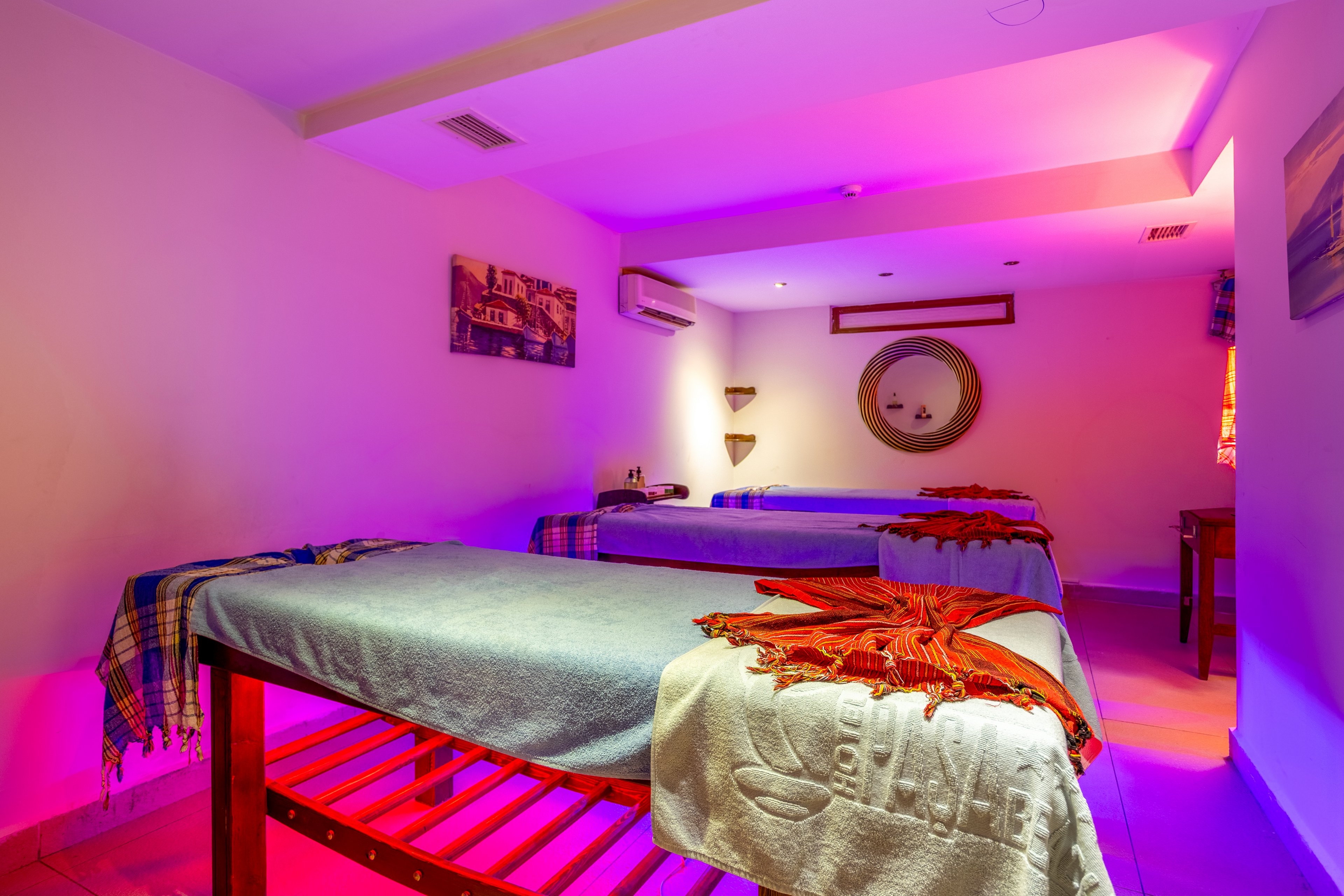A tranquil spa room features two massage tables neatly prepared with blankets and towels. Soft colored lighting in pink and purple tones creates a relaxing atmosphere. Wall art, a round decorative mirror, and small shelves add visual interest to the otherwise minimal space. The room appears ready for a couple or two guests to enjoy a massage session.