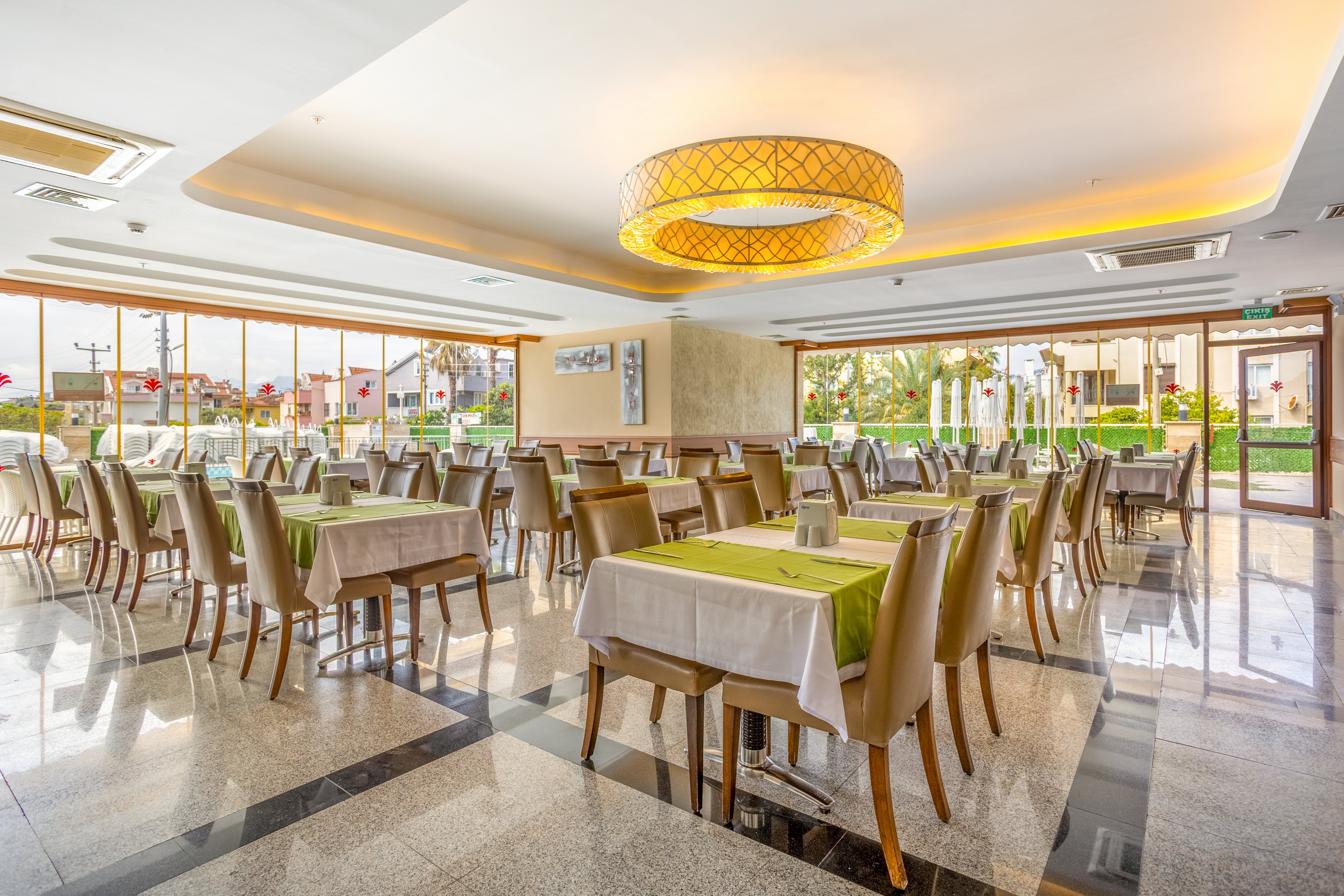 Spacious restaurant dining room with neatly arranged tables and chairs. Large windows let in abundant natural light and offer views of the outdoor garden area. Tables are covered with white cloths and green runners, creating a fresh, contemporary look. A circular ceiling light and polished floor add to the clean, modern atmosphere.