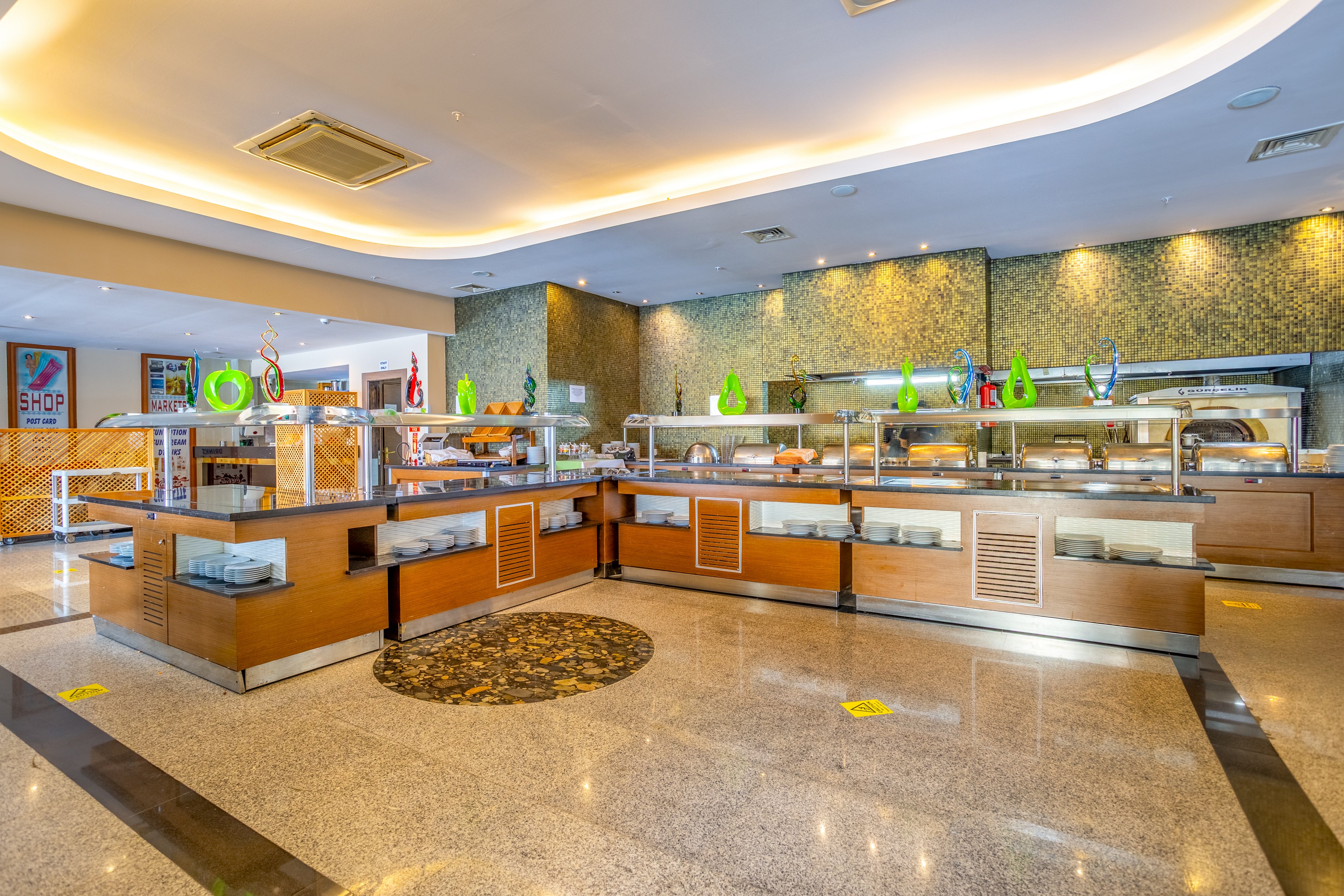 A modern self-service buffet counter stands in a spacious, well-lit dining hall. Wooden and stainless steel serving stations display covered trays, plates, and decorative glass vases. The polished stone floor reflects the warm ceiling lights and recessed lighting along the walls. No people are present, emphasizing the clean, organized layout and contemporary interior design.