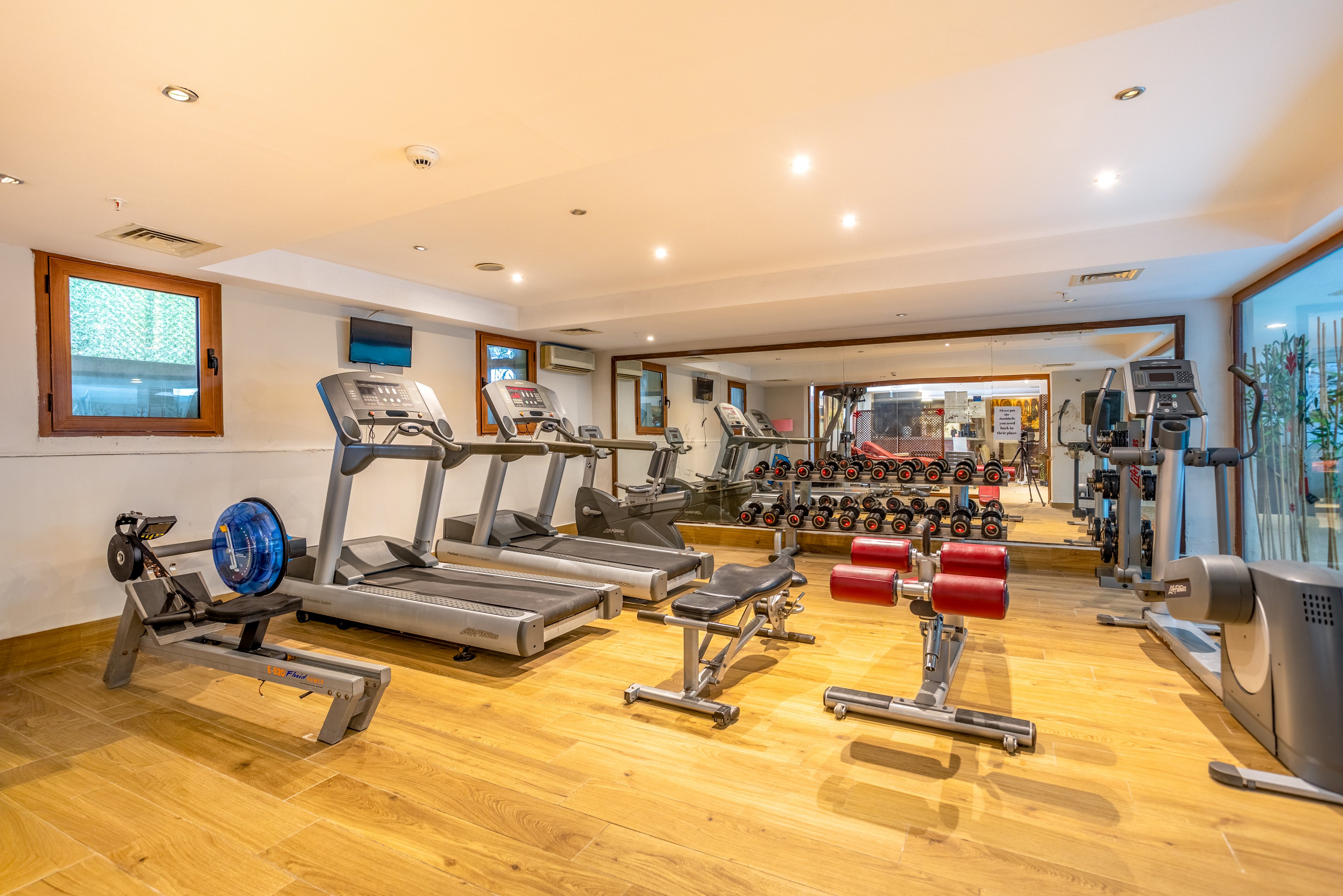 A bright, modern fitness room features several treadmills and other cardio machines on a wooden floor. Free weights and dumbbells are neatly arranged on racks in front of a large wall mirror. Strength-training benches and equipment are positioned throughout the space, creating a well-equipped workout area. Recessed ceiling lights and warm tones give the gym a clean and inviting atmosphere.