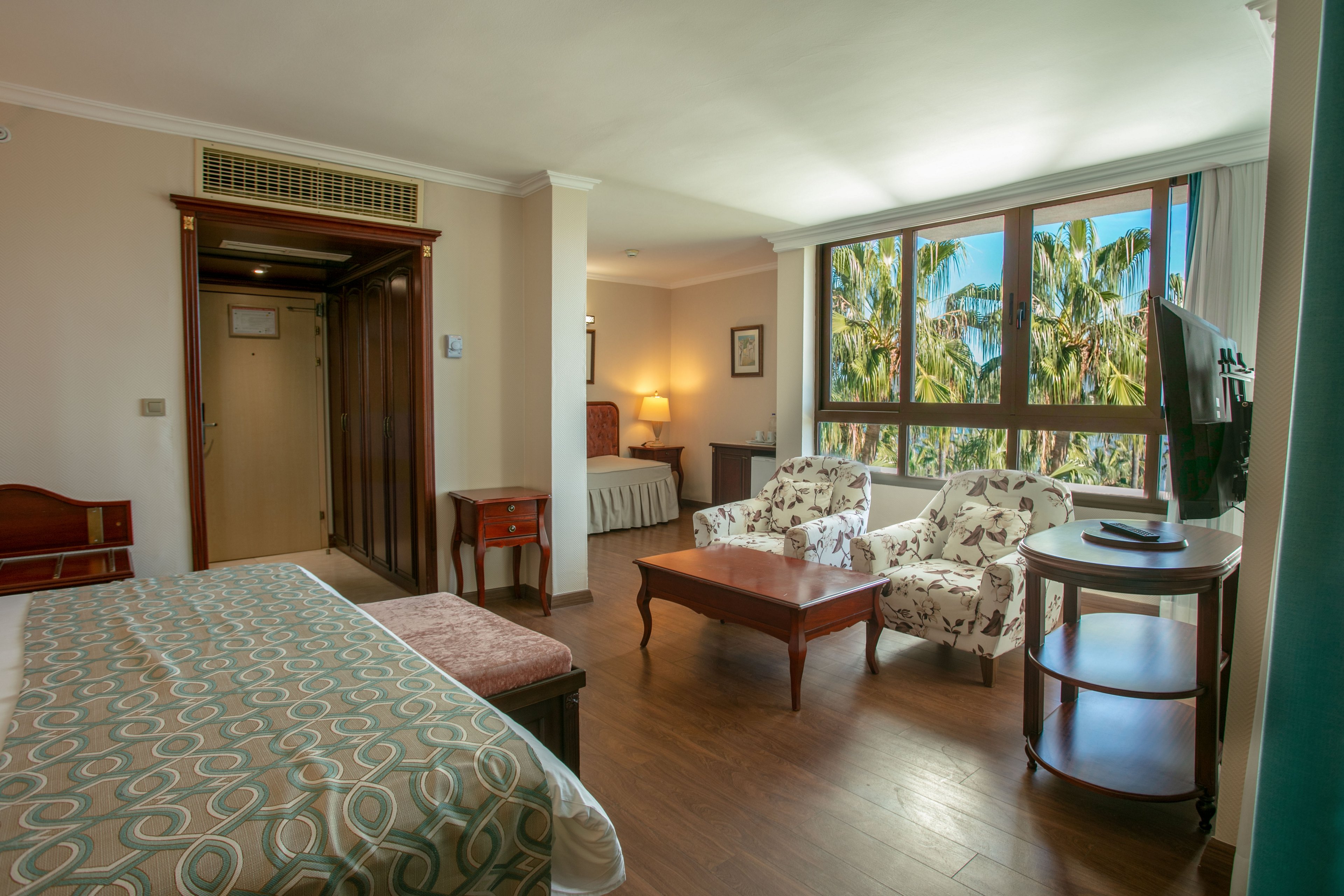 Spacious hotel room features a large bed and a cozy seating area with two patterned armchairs and a wooden coffee table. A flat-screen TV is positioned on a round stand near the window, facing the bed. Large windows let in abundant natural light and reveal lush green palm trees outside. Warm wooden flooring and soft lighting create a comfortable, inviting atmosphere.