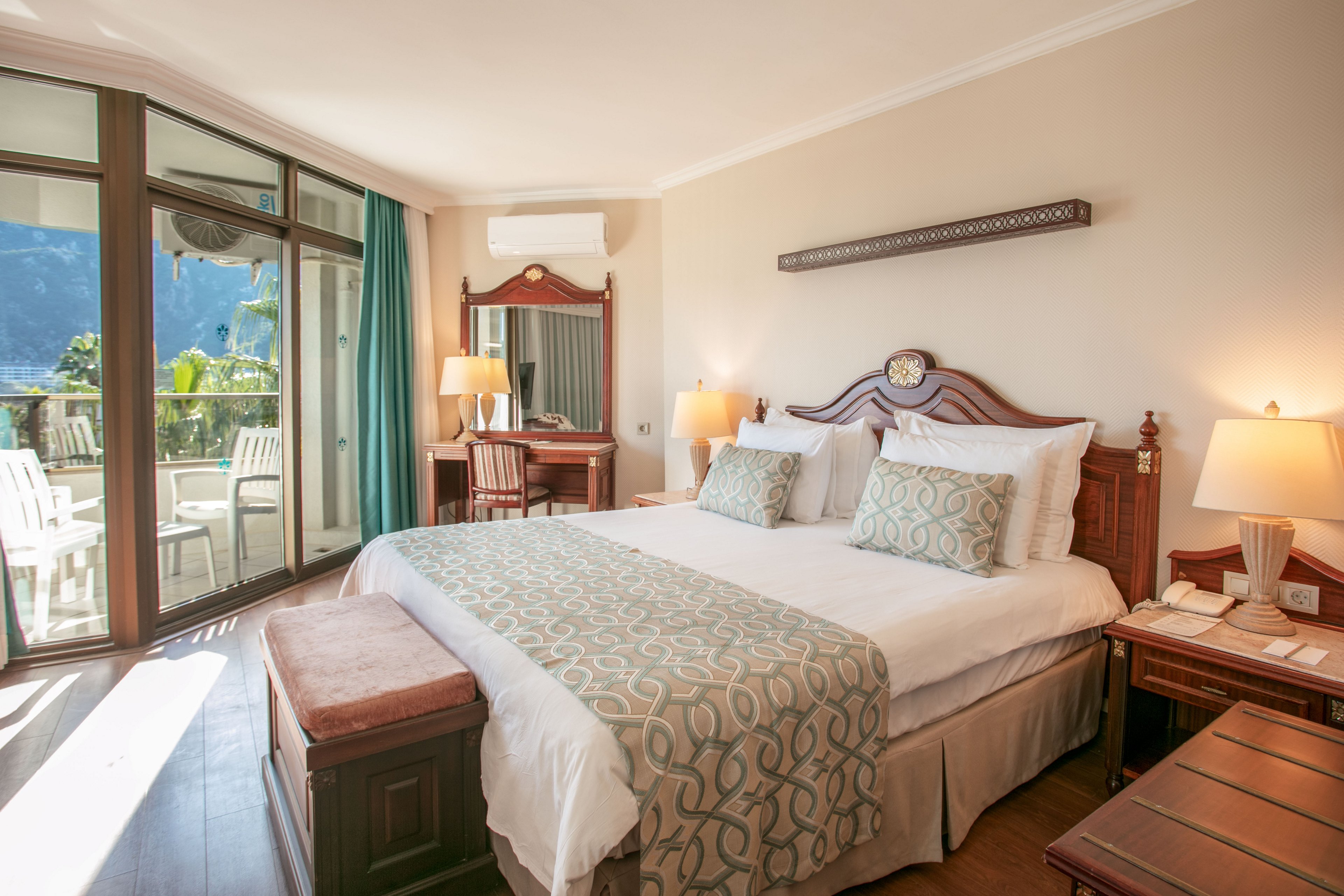 A well-lit hotel bedroom features a large double bed with patterned pillows and a matching runner. Wooden furniture, including bedside tables, a bench, and a vanity desk with mirror, adds a classic touch to the room. A sliding glass door opens to a balcony with white chairs and a scenic mountain and water view. Warm lighting from a bedside lamp and neutral wall colors create a cozy, inviting atmosphere.