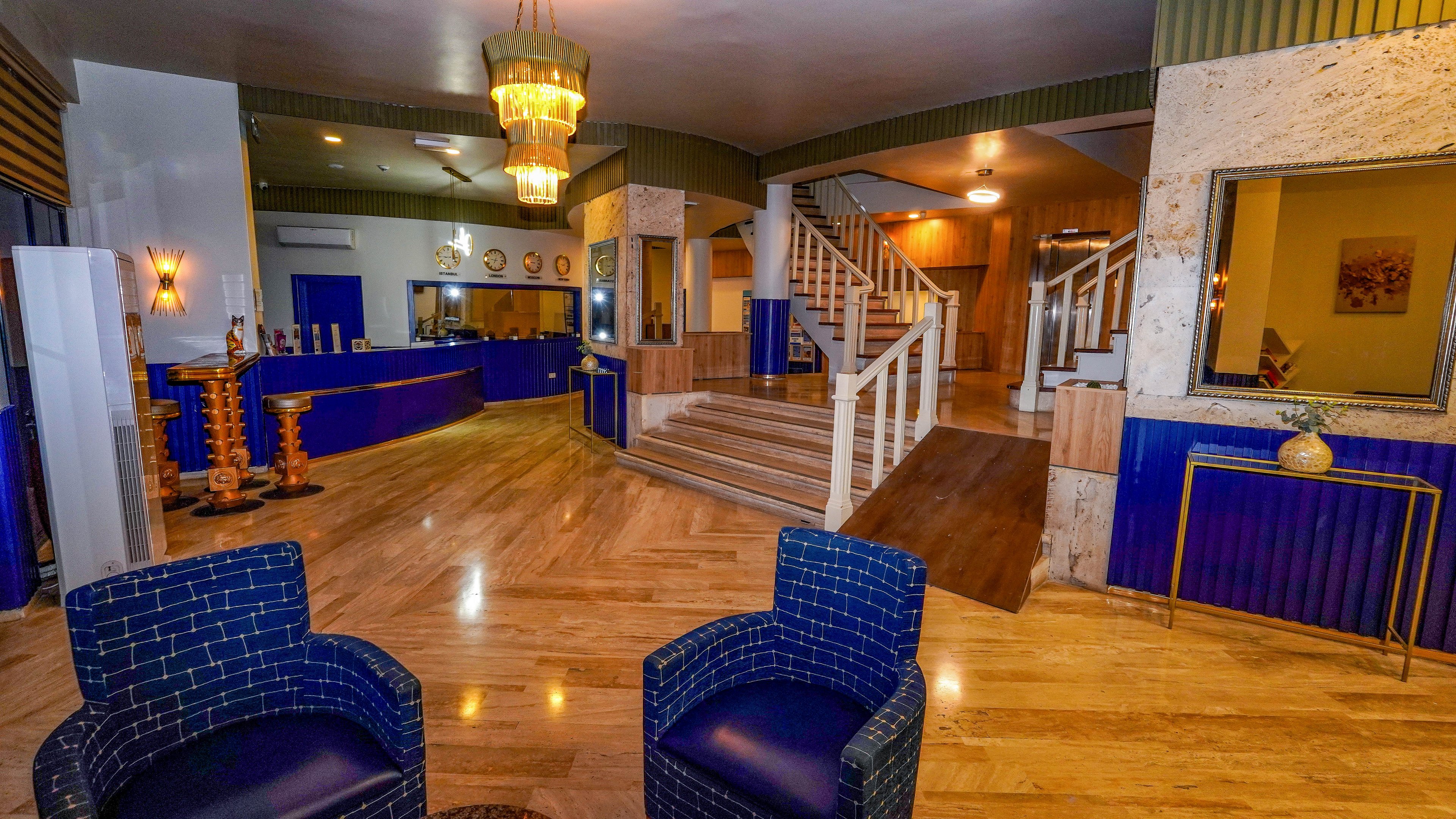 A contemporary hotel lobby features a spacious reception area with bold blue counters and seating. Two patterned blue armchairs sit in the foreground on polished wooden floors, leading the eye toward a central staircase. Warm lighting from chandeliers and wall sconces creates a welcoming atmosphere, highlighting the mix of wood, stone, and metal finishes. Decorative accents and framed artwork add visual interest throughout the space.