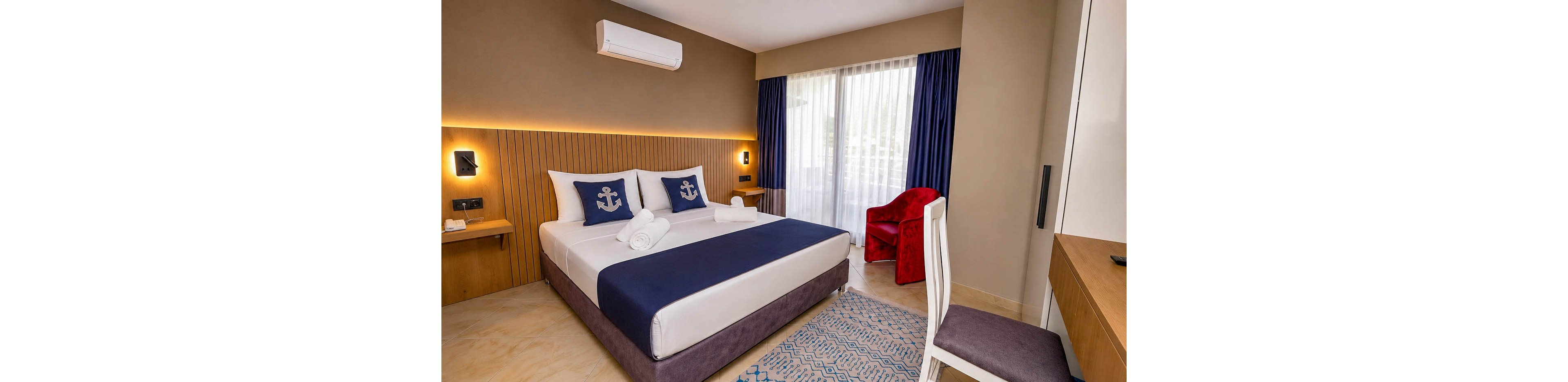 A contemporary hotel bedroom features a neatly made double bed with navy blue bedding and anchor-themed pillows. The room includes a wooden headboard wall with integrated lighting, a desk with a white chair, and a red armchair near the window. Large glass doors with sheer curtains and dark blue drapes allow natural light into the space. A patterned rug and neutral-toned walls contribute to the clean, modern aesthetic.