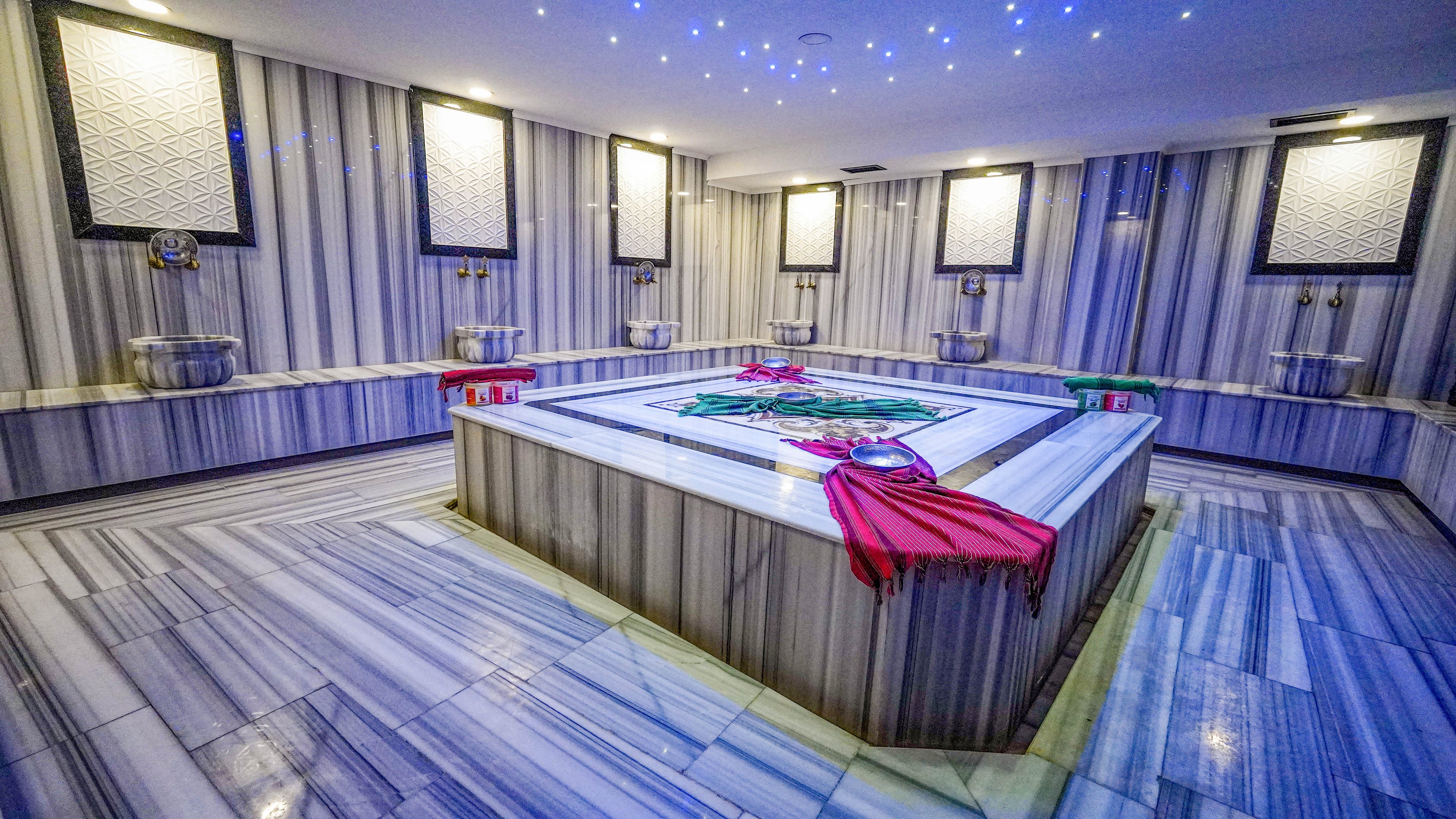 Spacious Turkish hammam with a large central marble platform dominates the scene. Striped gray and white marble covers the floors and walls, creating a clean and elegant spa environment. Colorful towels in red and green are draped over the heated stone slab, adding contrast to the cool-toned room. Multiple basins and wall-mounted faucets line the perimeter, while recessed ceiling lights and star-like blue LEDs enhance the relaxing atmosphere.