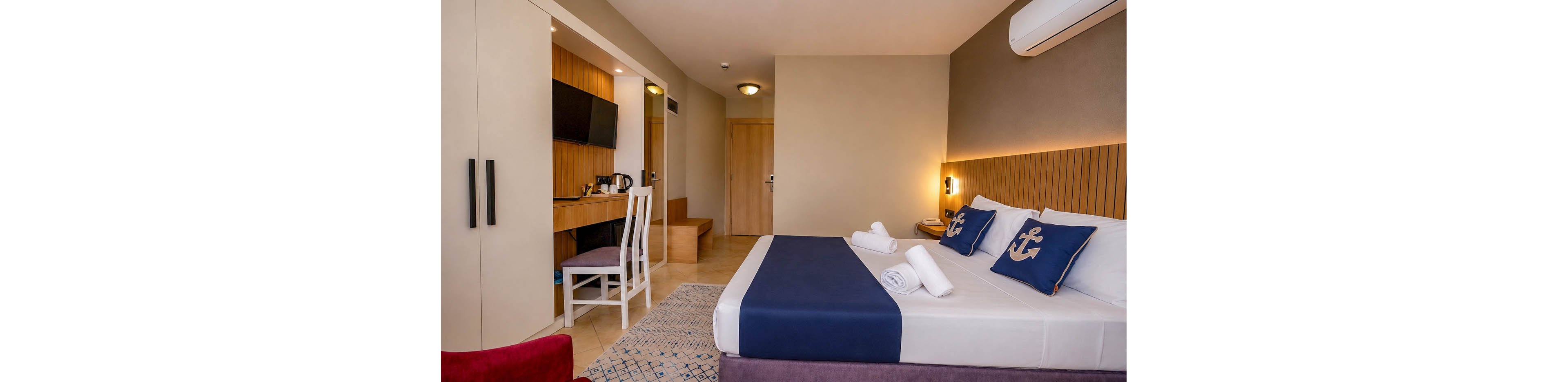 A contemporary hotel room features a neatly made double bed with navy bedding and anchor-themed pillows. The space includes a wooden desk with a flat-screen television, electric kettle, and white chair. Warm neutral walls, recessed lighting, and a patterned rug create a cozy, minimalist atmosphere. A doorway and built-in wardrobe are visible in the background, emphasizing the room’s functional layout.