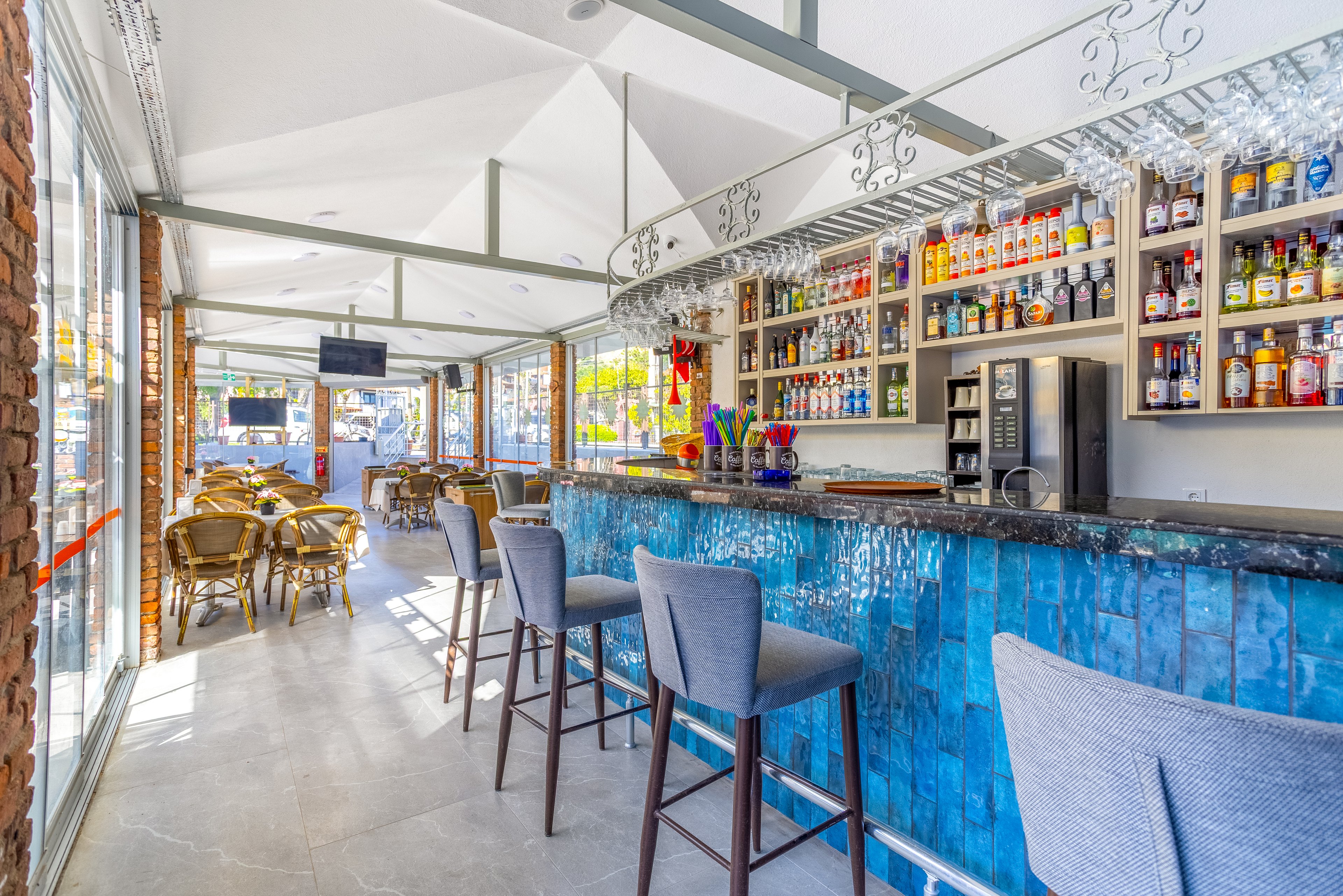A contemporary bar interior features a long counter clad in vibrant blue tiles. Shelves behind the bar are filled with assorted liquor bottles and glassware, creating a colorful backdrop. High bar stools line the counter, while wicker chairs and tables are arranged along the sunlit windows. Large windows and glass doors let in abundant natural light, giving the space an airy, inviting feel.