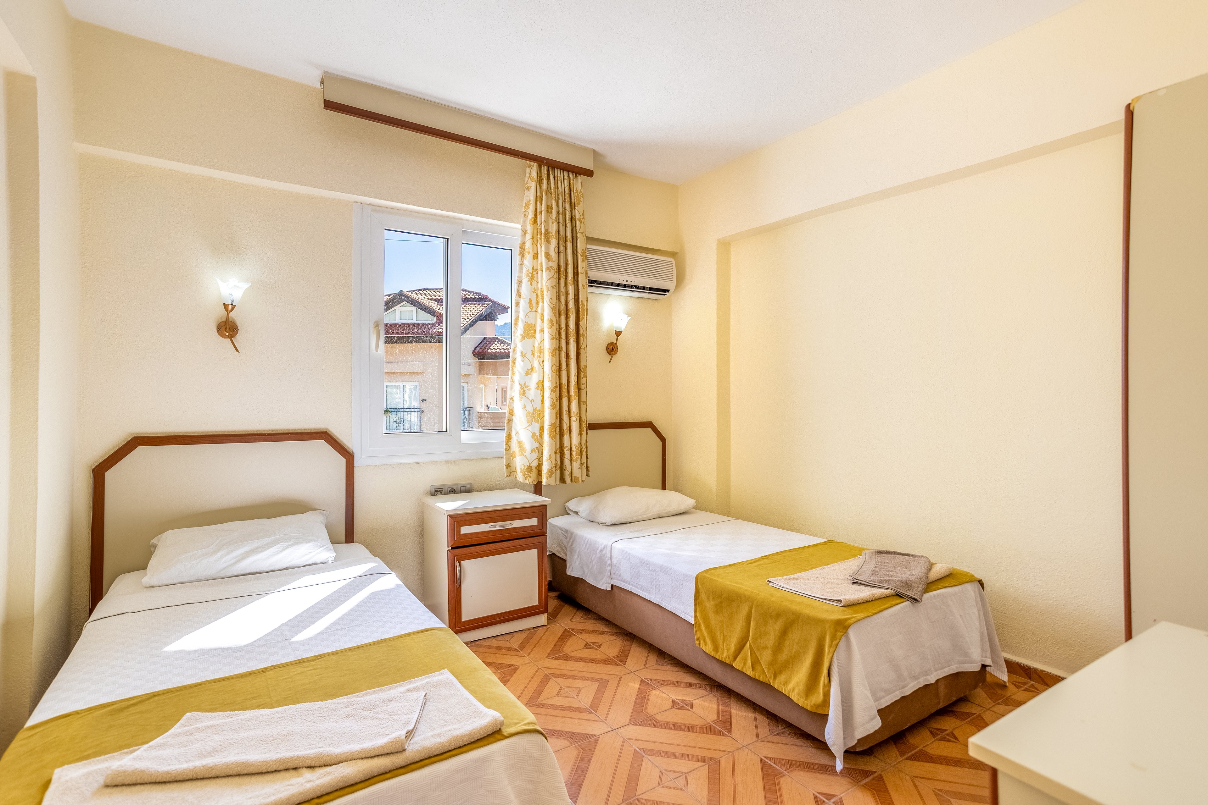 A small, tidy bedroom features two single beds with white linens and yellow bed runners. The room is warmly lit by wall sconces and natural light from a window with patterned yellow curtains. An air conditioning unit is mounted above the window, and a small nightstand sits between the beds. The light-colored walls and patterned wooden floor create a cozy, inviting atmosphere.