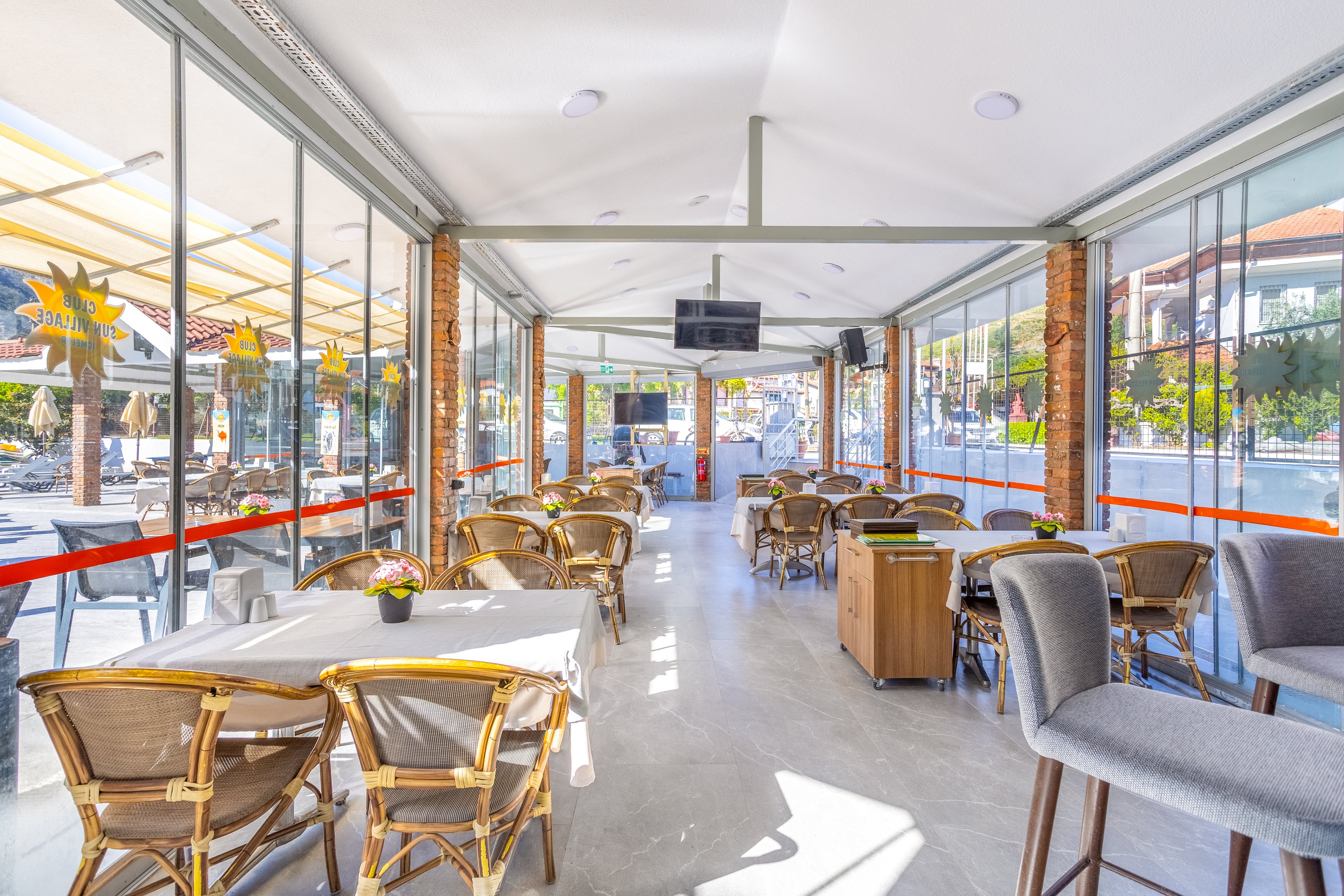 A bright modern cafe interior features wicker chairs and neatly arranged tables inside a glass-walled space. Large windows on both sides provide views of the outdoor seating area and surrounding buildings. The room is flooded with natural light, highlighting neutral tones and clean contemporary lines. Ceiling-mounted televisions and decorative plants add to the relaxed, casual dining atmosphere.