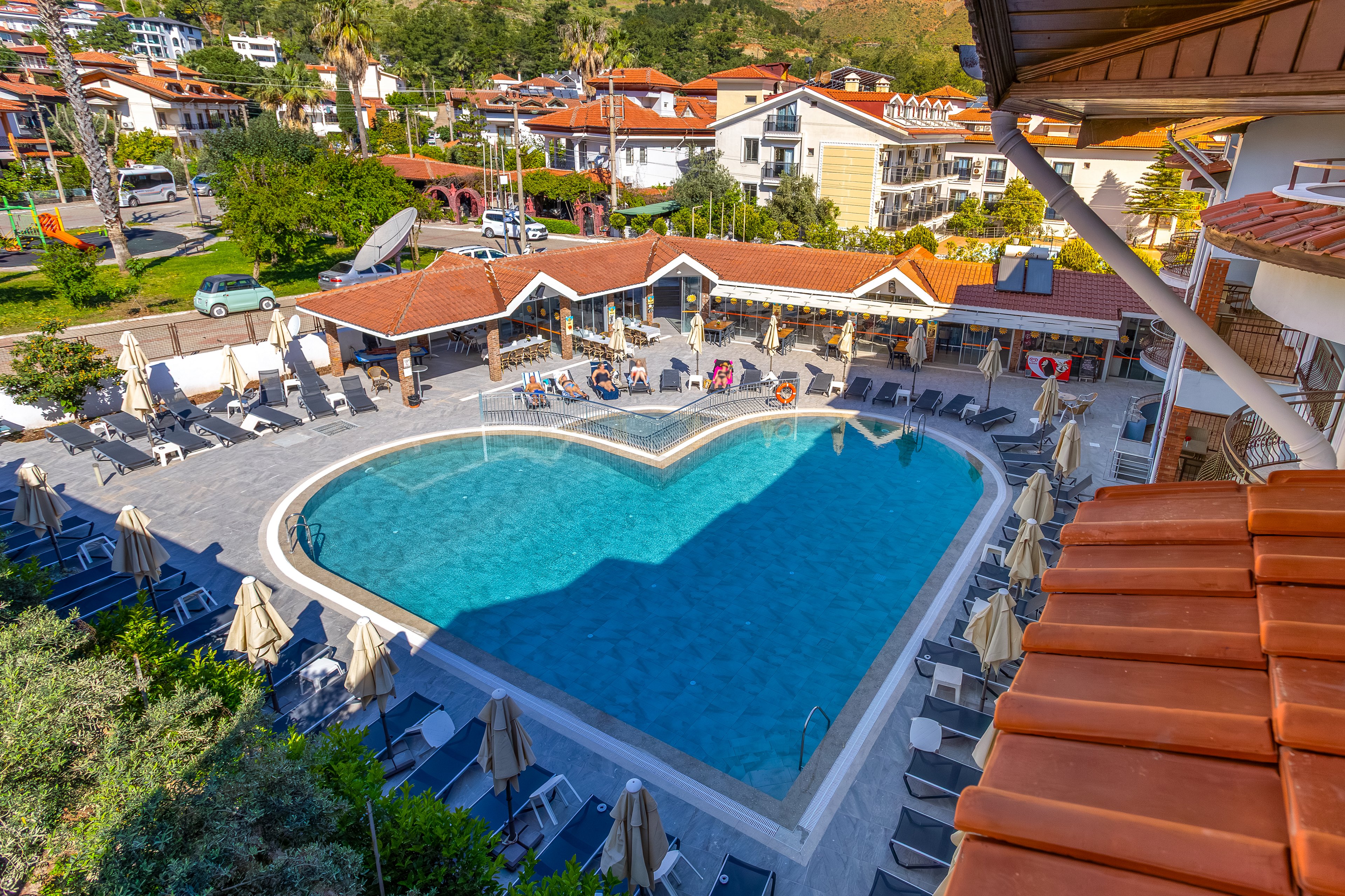 A large heart-shaped swimming pool is the central focus of this resort complex. Numerous sun loungers and umbrellas surround the pool, creating a relaxed vacation atmosphere. The pool area is bordered by low buildings with red-tiled roofs, likely housing bars, restaurants, or guest facilities. In the background, residential-style structures, parked cars, and green hills complete the scenic holiday setting.
