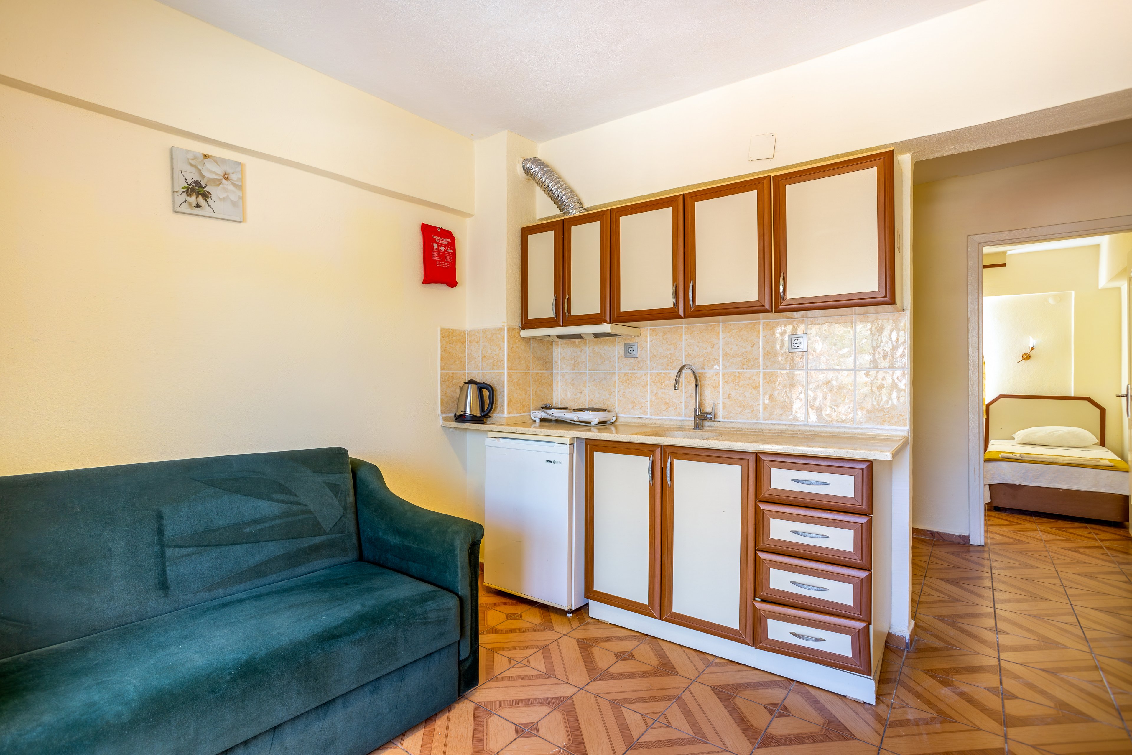 A small studio apartment features a compact kitchenette with wooden cabinets and cream-colored doors. The kitchen area includes a sink, stovetop, kettle, and a mini refrigerator beneath tiled walls. A green upholstered sofa sits opposite the kitchenette on patterned wooden flooring. In the background, an open doorway reveals a simple bedroom with a single bed and soft lighting. The overall space appears bright, tidy, and functional for short-term stays.