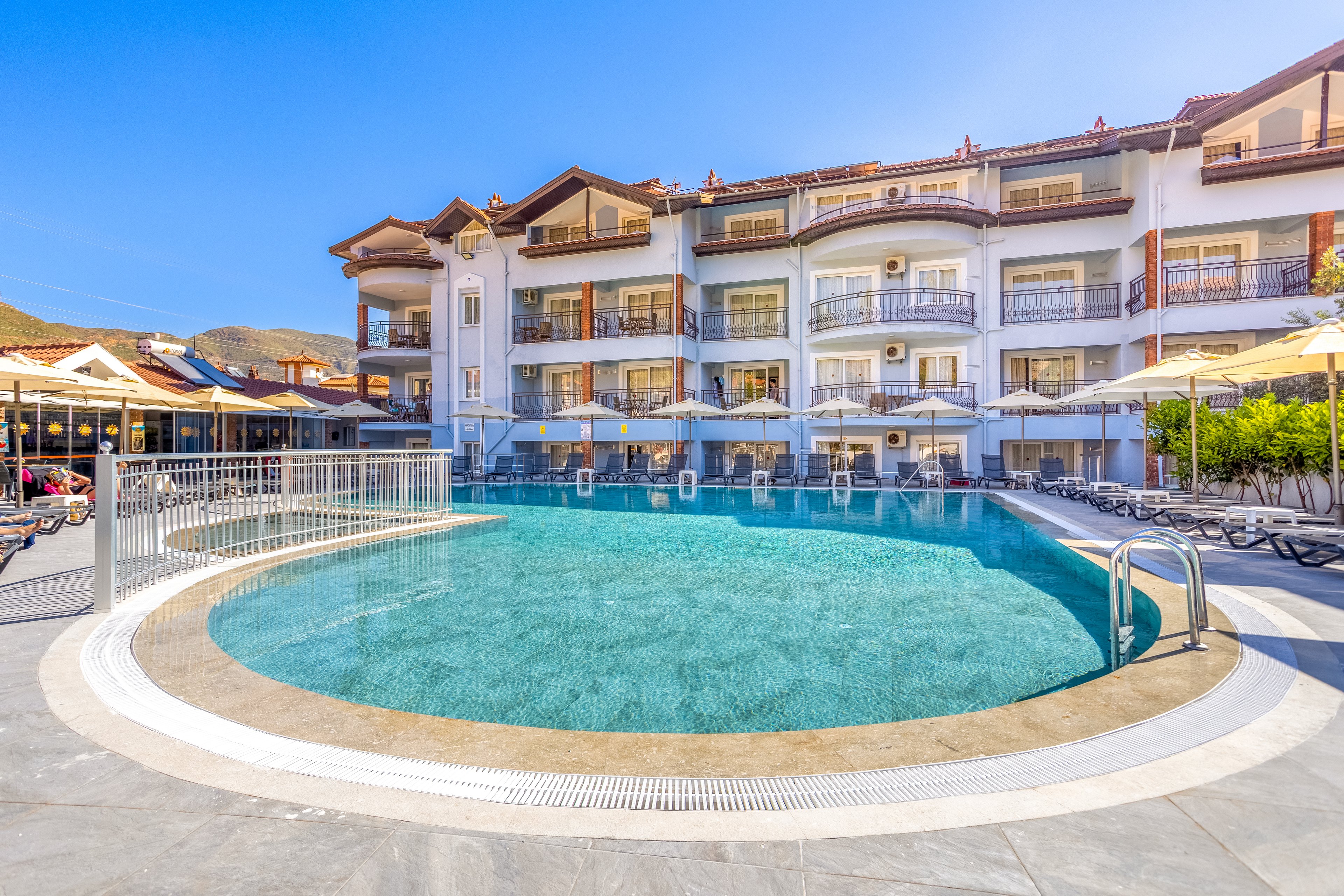 A bright resort hotel features a large curved outdoor swimming pool in the foreground. The multi-story building has balconies with railings and sunshades, facing the pool area. Numerous sun loungers and umbrellas line the pool deck, suggesting a relaxing vacation setting. Clear blue skies and distant hills enhance the warm, sunny atmosphere of the scene.