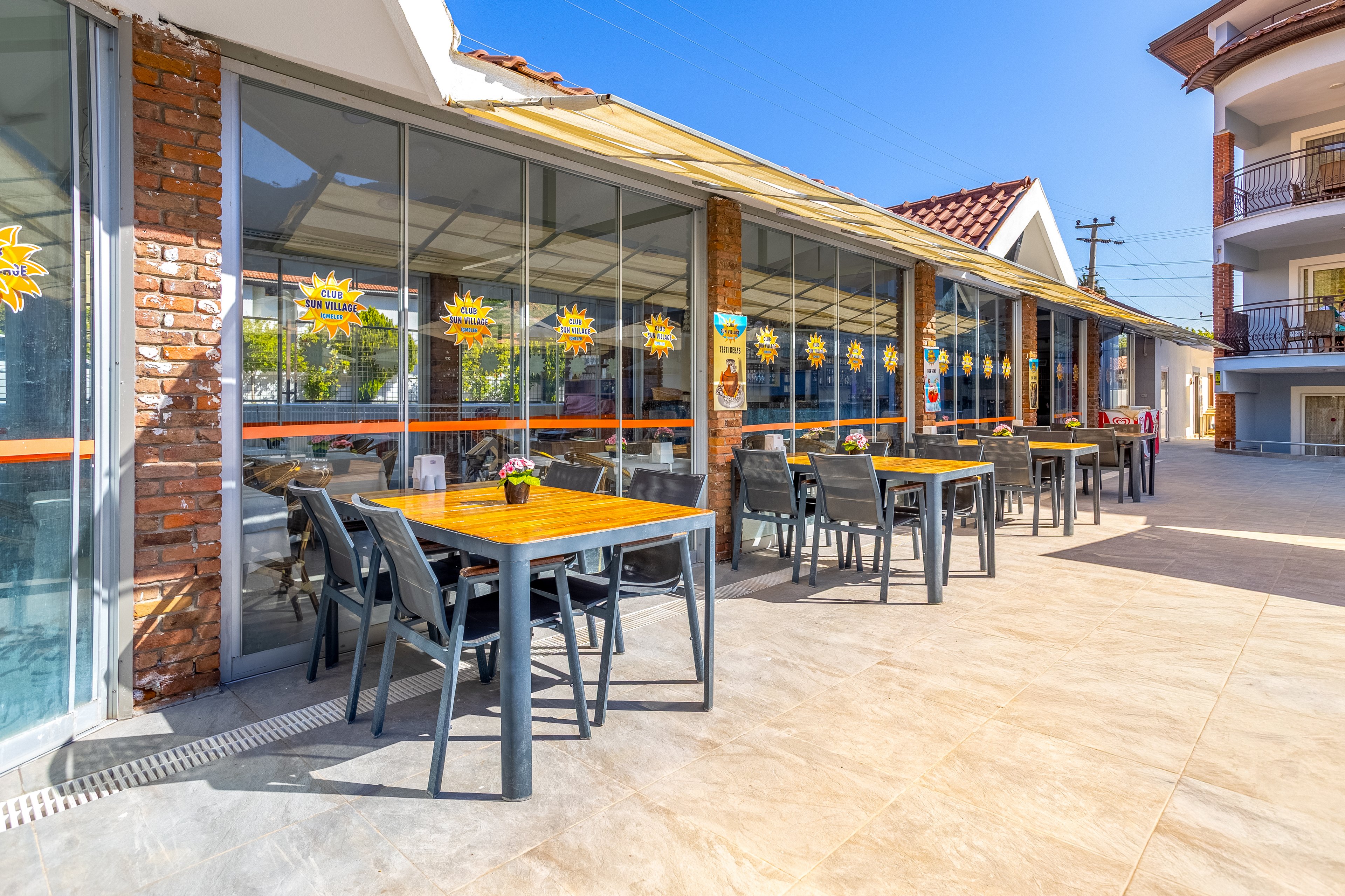 An outdoor cafe terrace features several wooden tables with dark chairs neatly arranged along a glass-fronted building. The setting appears to be part of a hotel or resort complex, with balconies and additional structures visible in the background. Bright sunlight and clear blue skies create a warm, inviting atmosphere. Decorative sun-shaped decals on the windows add a cheerful visual element to the scene.